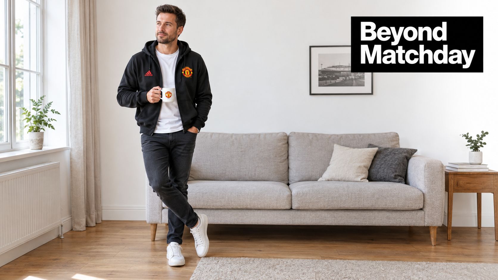 A man wearing a black Manchester United hoodie and jeans stands in a bright, modern living room.