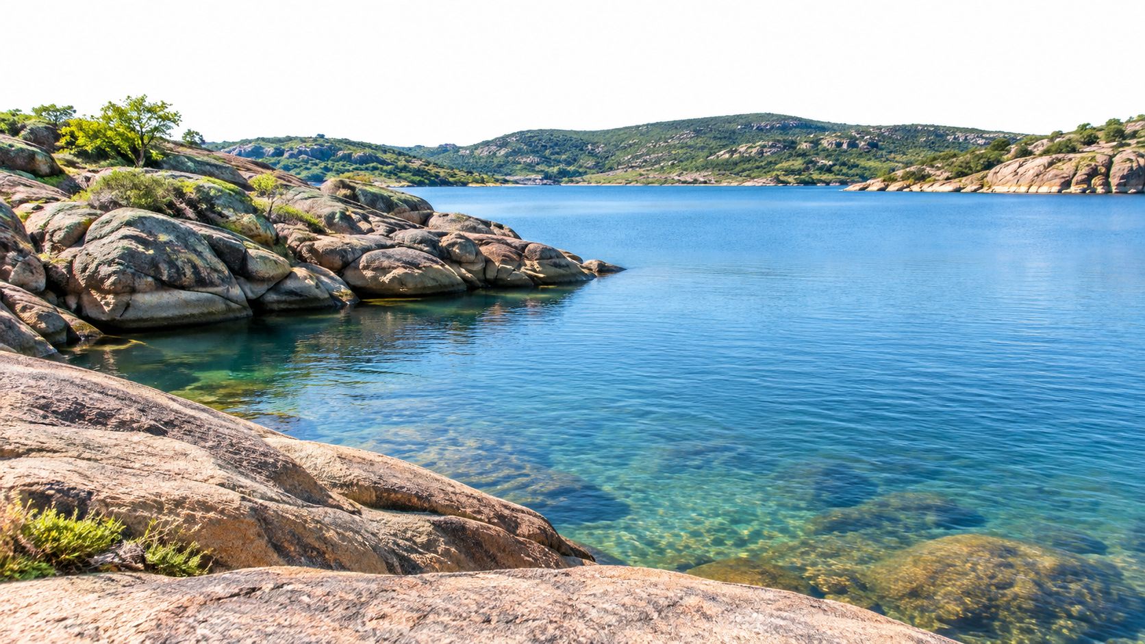 A serene and crystal clear blue lake surrounded by rocky shorelines and distant lush green rolling hills.