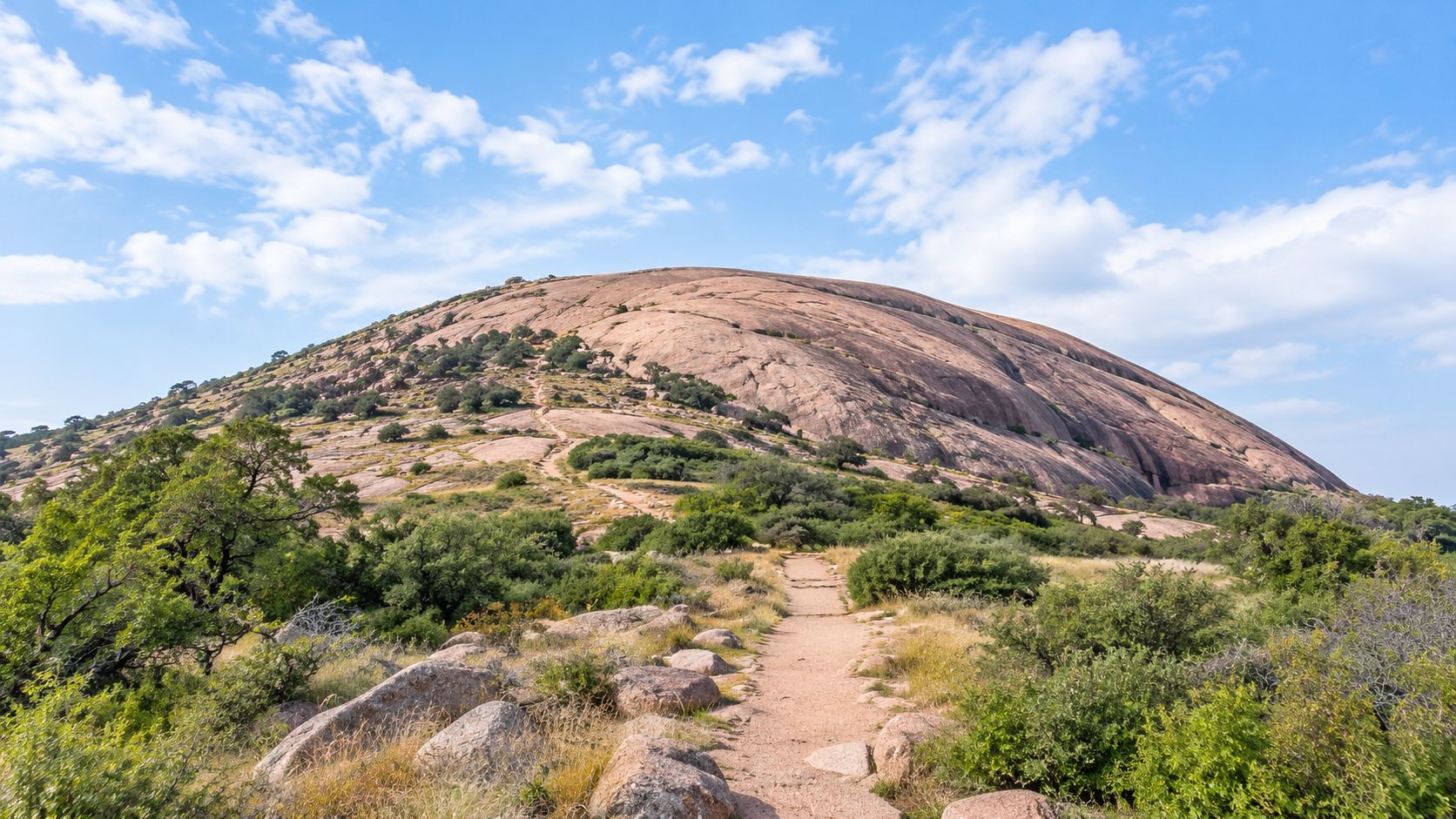 A dirt hiking trail leads toward the massive granite dome of Enchanted Rock in a Texas state park.