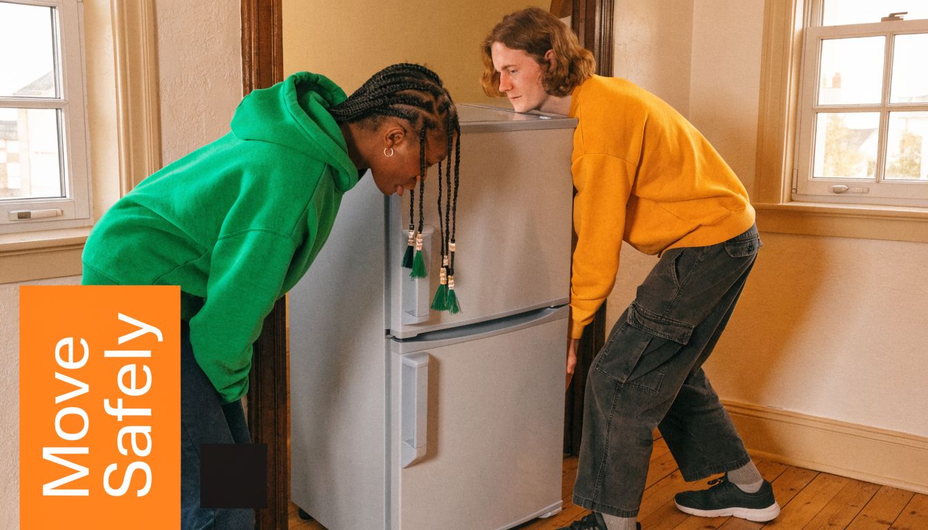 Two young people working together to carefully move a light grey fridge freezer through a doorway.
