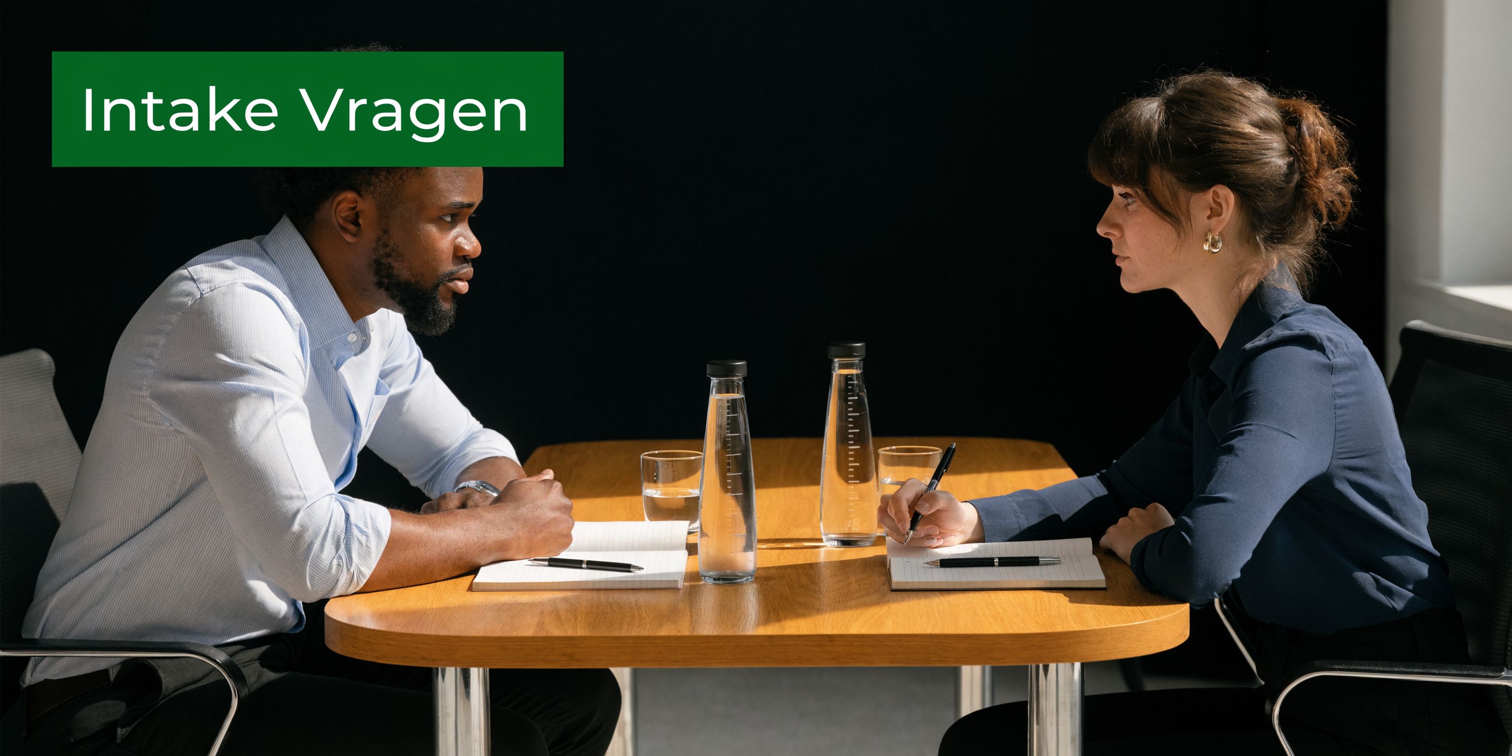 Een man en een vrouw zitten tegenover elkaar aan een houten tafel tijdens een professioneel intakegesprek