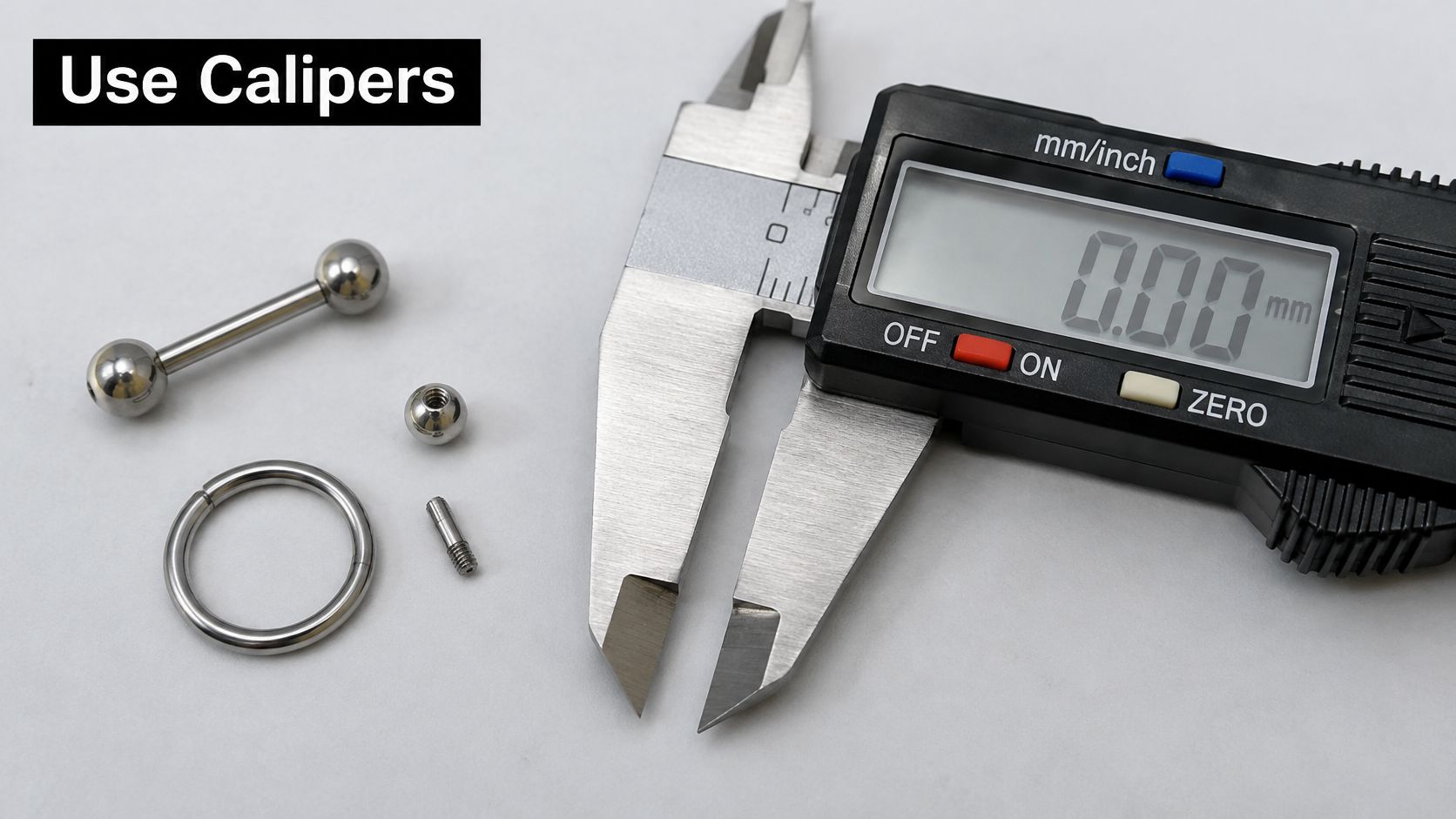 A pair of digital calipers lying on a white surface next to various metal body jewelry pieces.