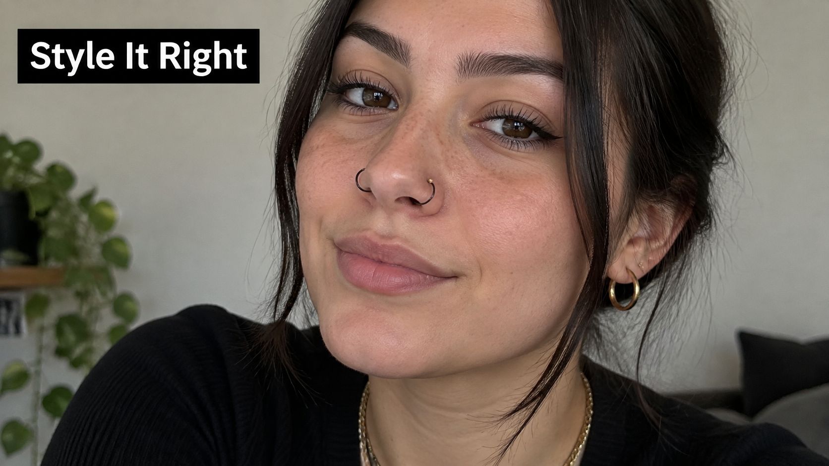 A young woman smiling, featuring a black hoop nose piercing and stylish gold hoop earrings.