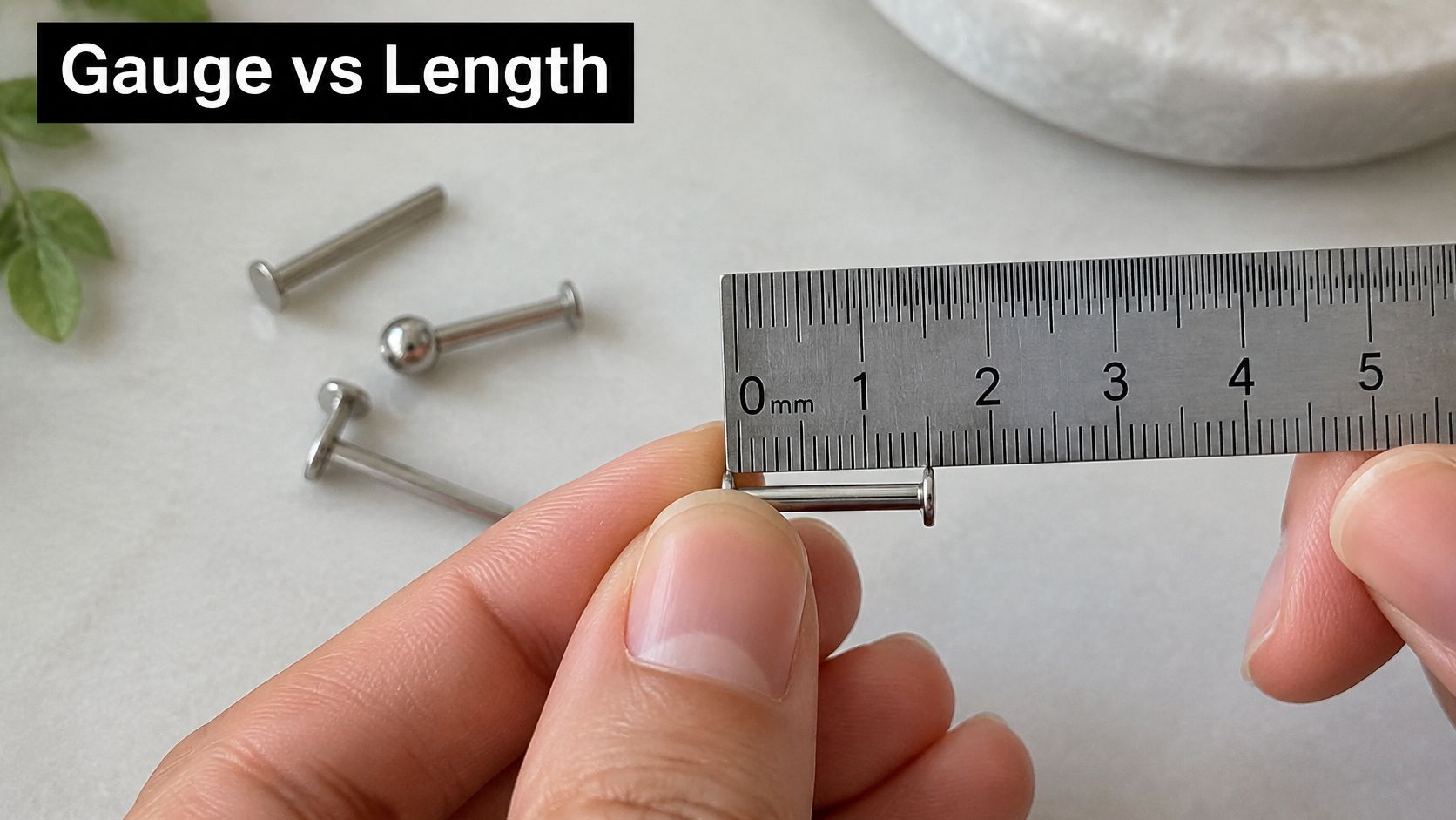 A hand holding a metal piercing stud against a metal ruler to measure its length in millimeters.