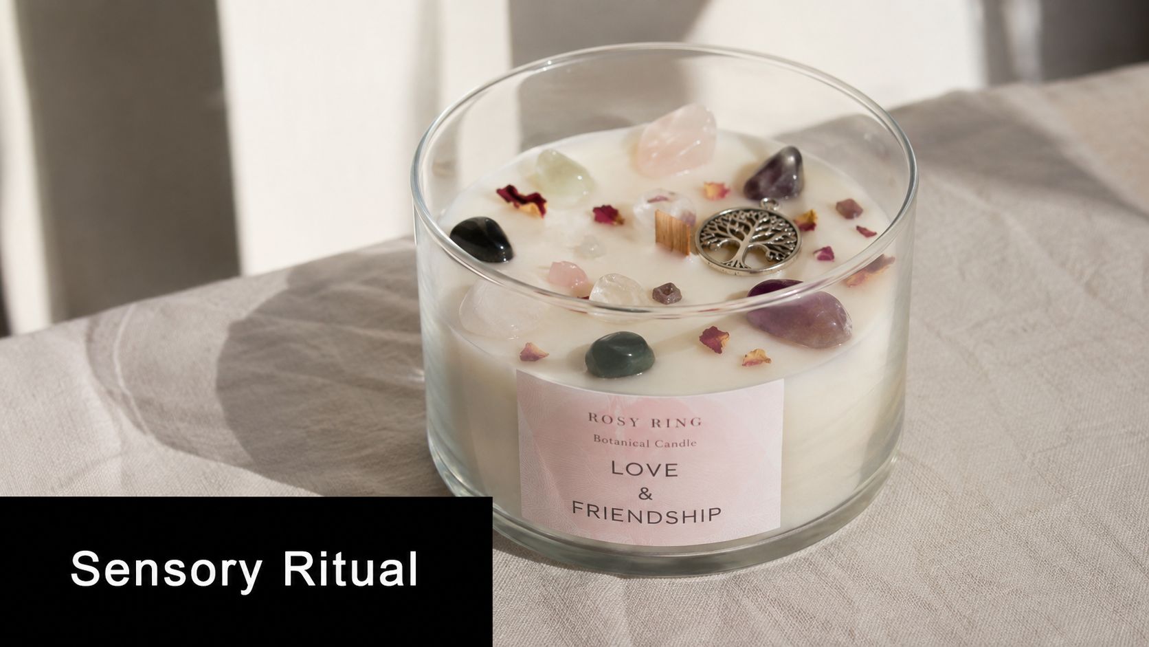 A glass candle jar labeled Rosy Ring Love & Friendship decorated with various crystals and a tree charm.