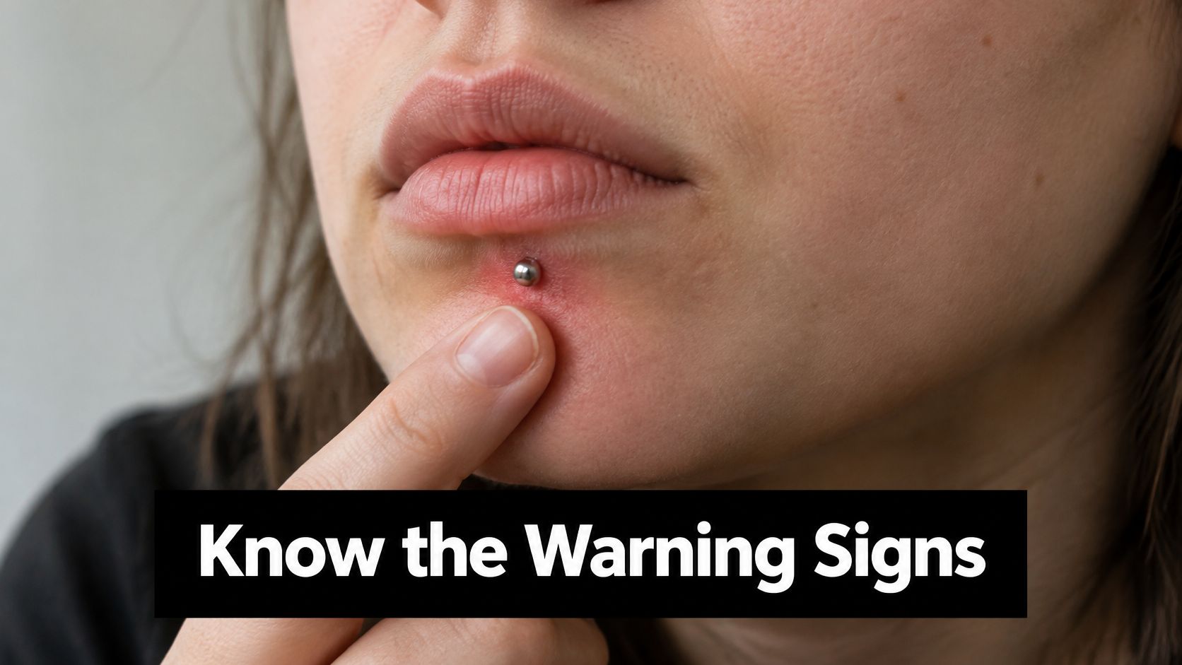 A close-up of a person touching an irritated, red labret piercing on their lower lip.