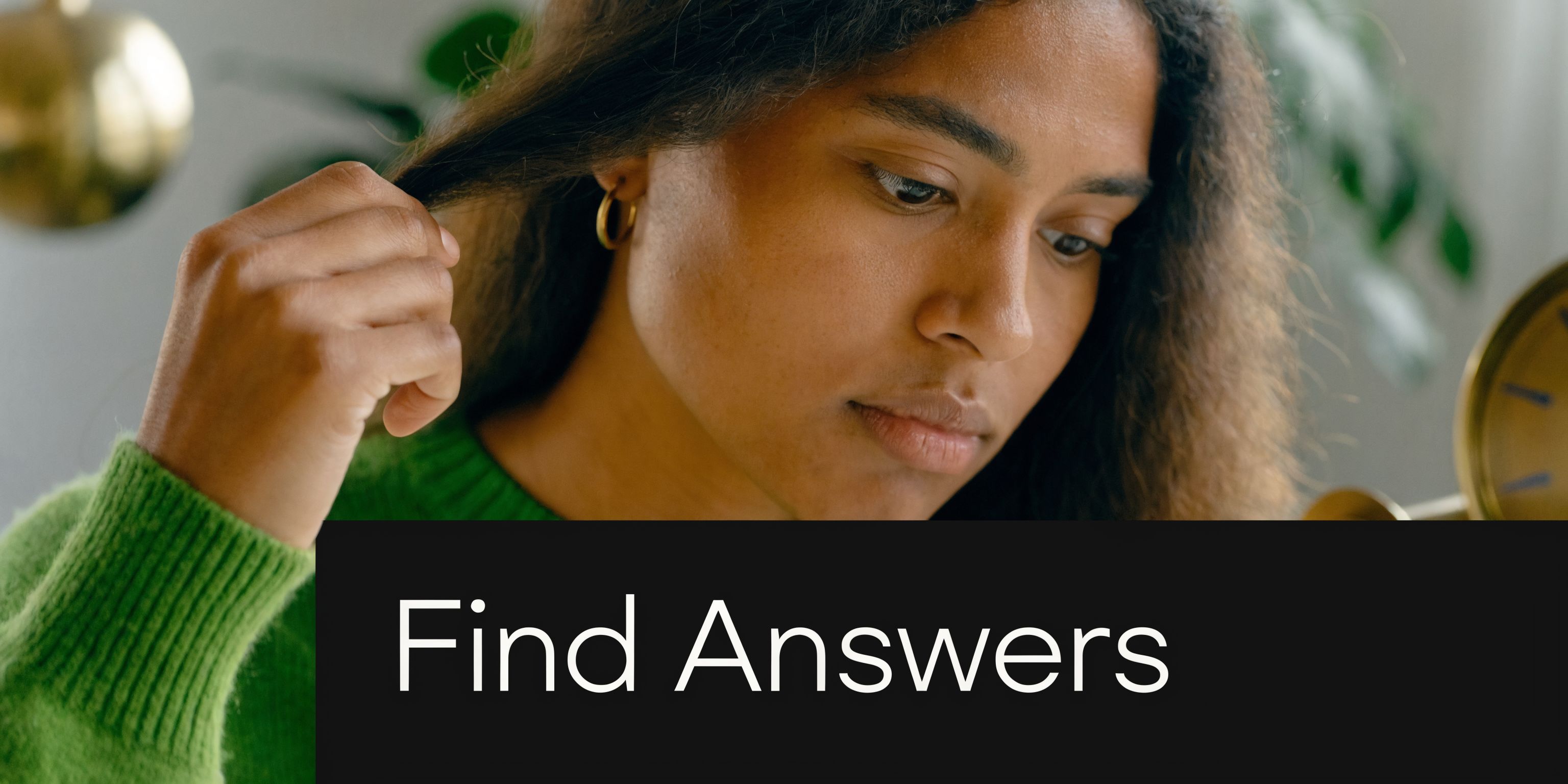 A focused young woman in a green sweater examines a strand of her curly hair closely.