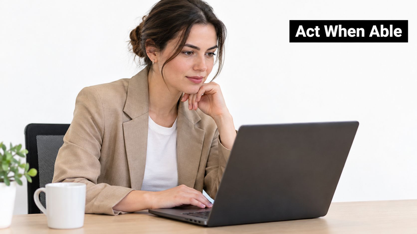 A professional woman in a tan blazer sitting at a desk and using a laptop computer.
