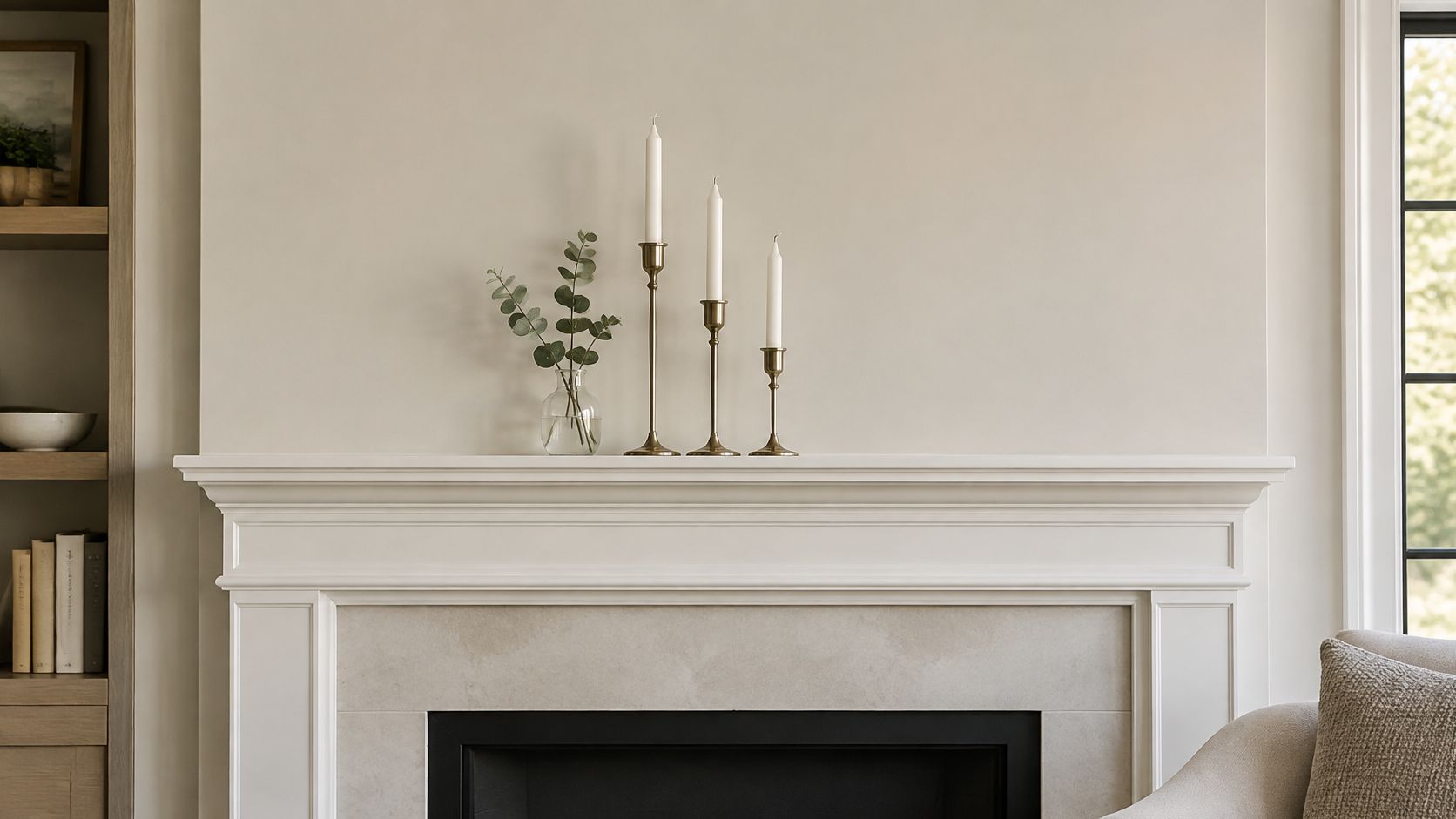 A modern fireplace mantel decorated with tall brass candle holders and a small glass vase with eucalyptus branches.