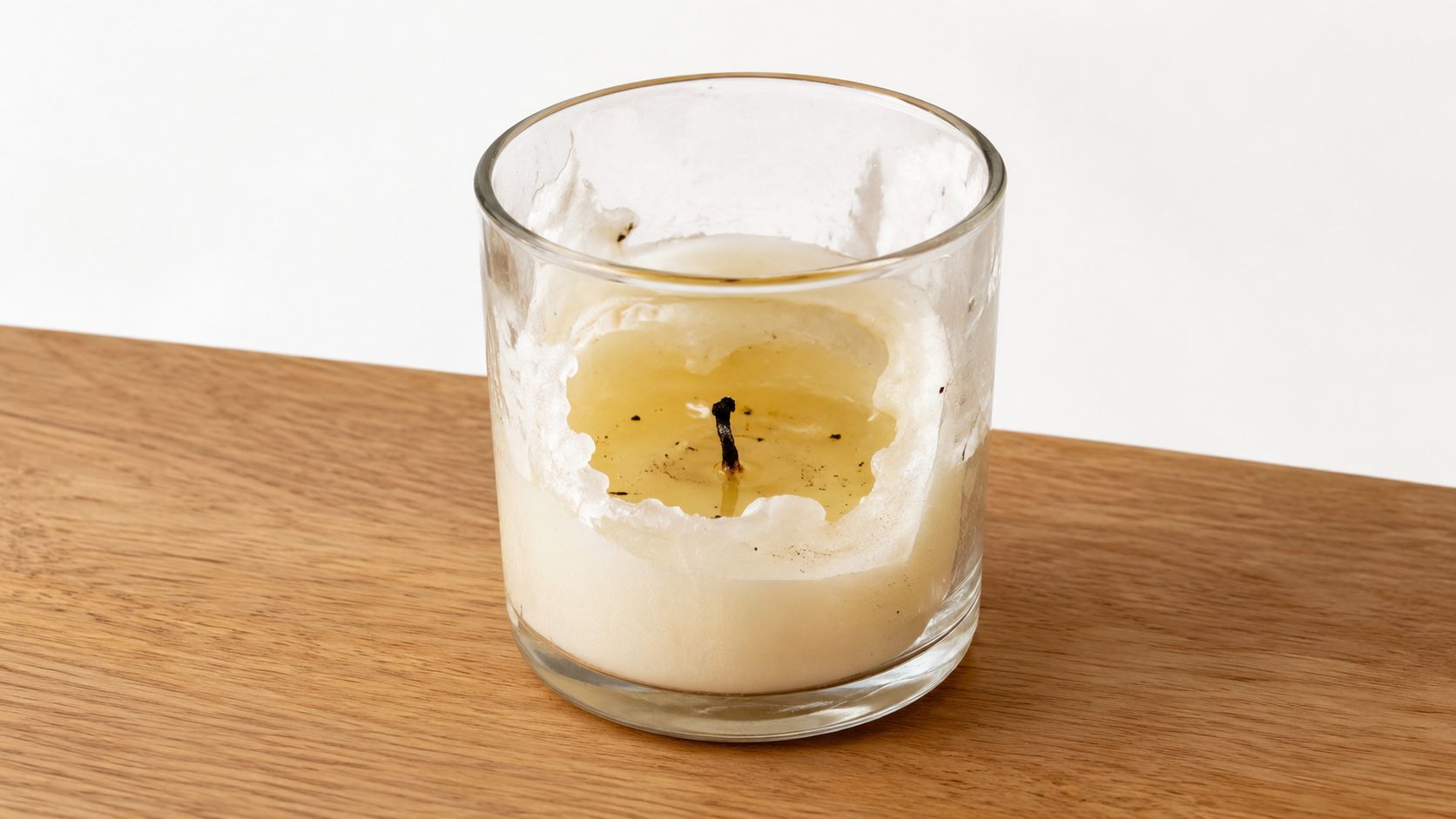 A half-used candle in a glass container sitting on a light wooden surface against a plain background.