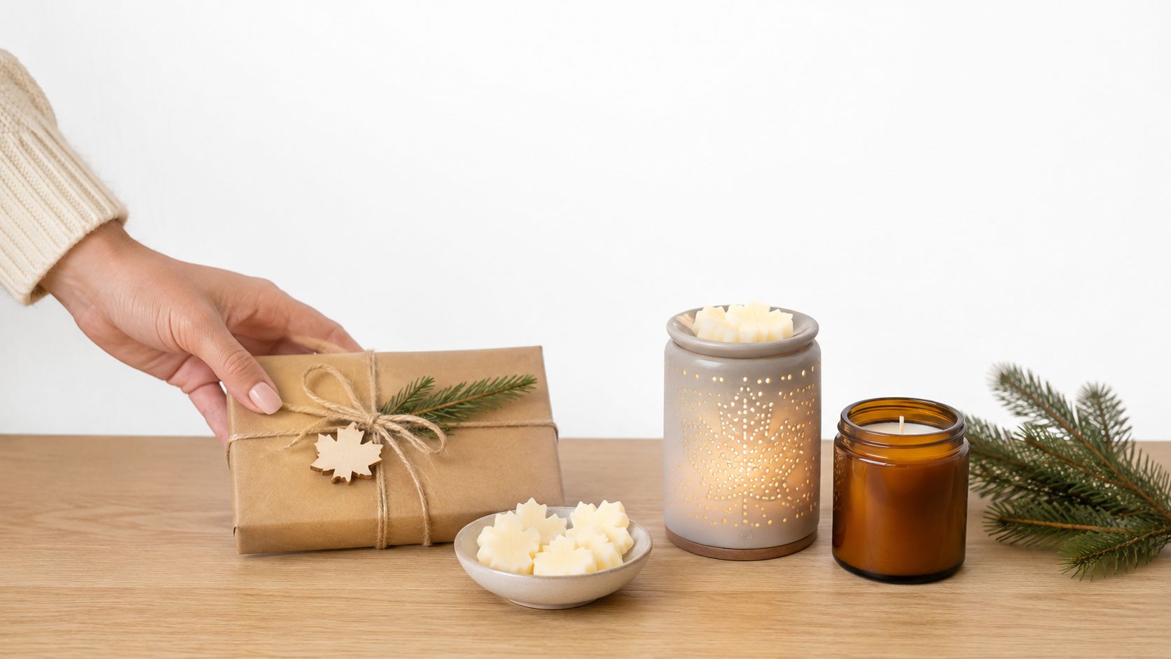 A hand placing a wrapped gift next to maple leaf wax melts, a warmer, and a candle.