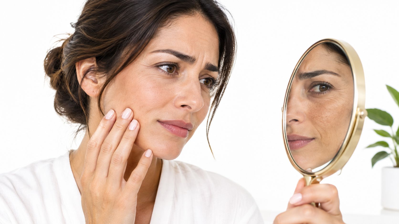 A woman examining her skin in a hand mirror, concerned about signs of aging and skin texture.