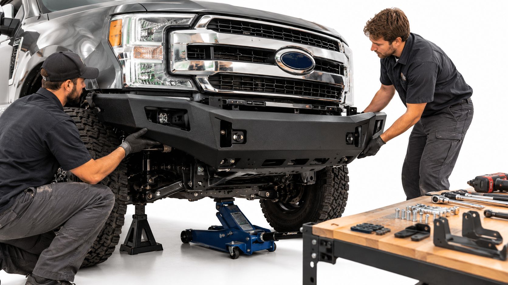 Two automotive technicians installing a heavy-duty steel off-road bumper onto a gray Ford pickup truck.