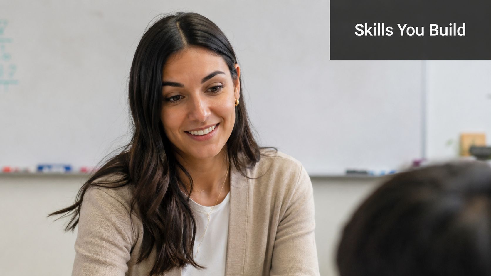 A friendly teacher smiling while providing instruction and support to a student in a classroom setting.