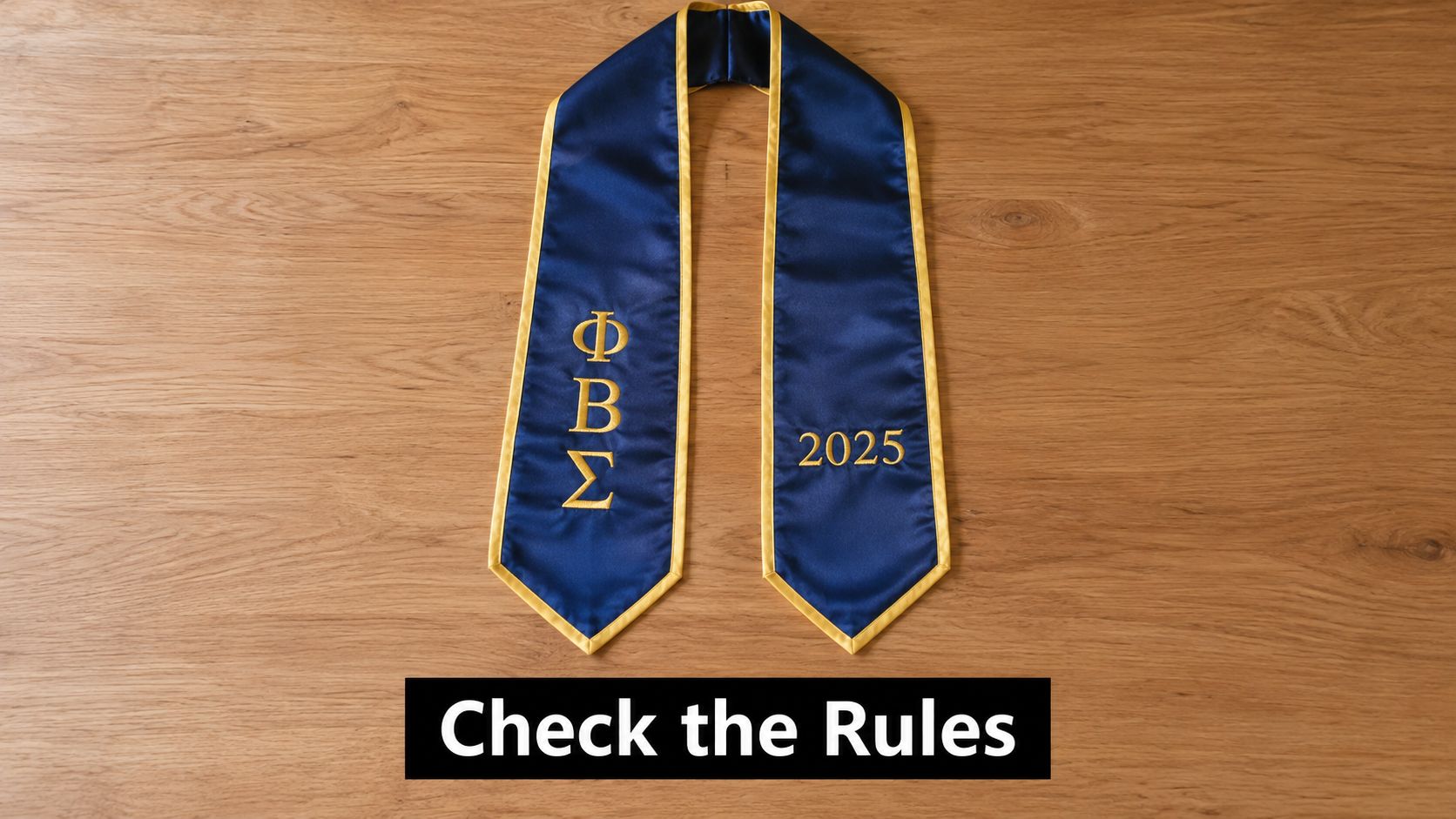 A blue graduation sash with gold Greek letters and the year 2025 on a wooden background.