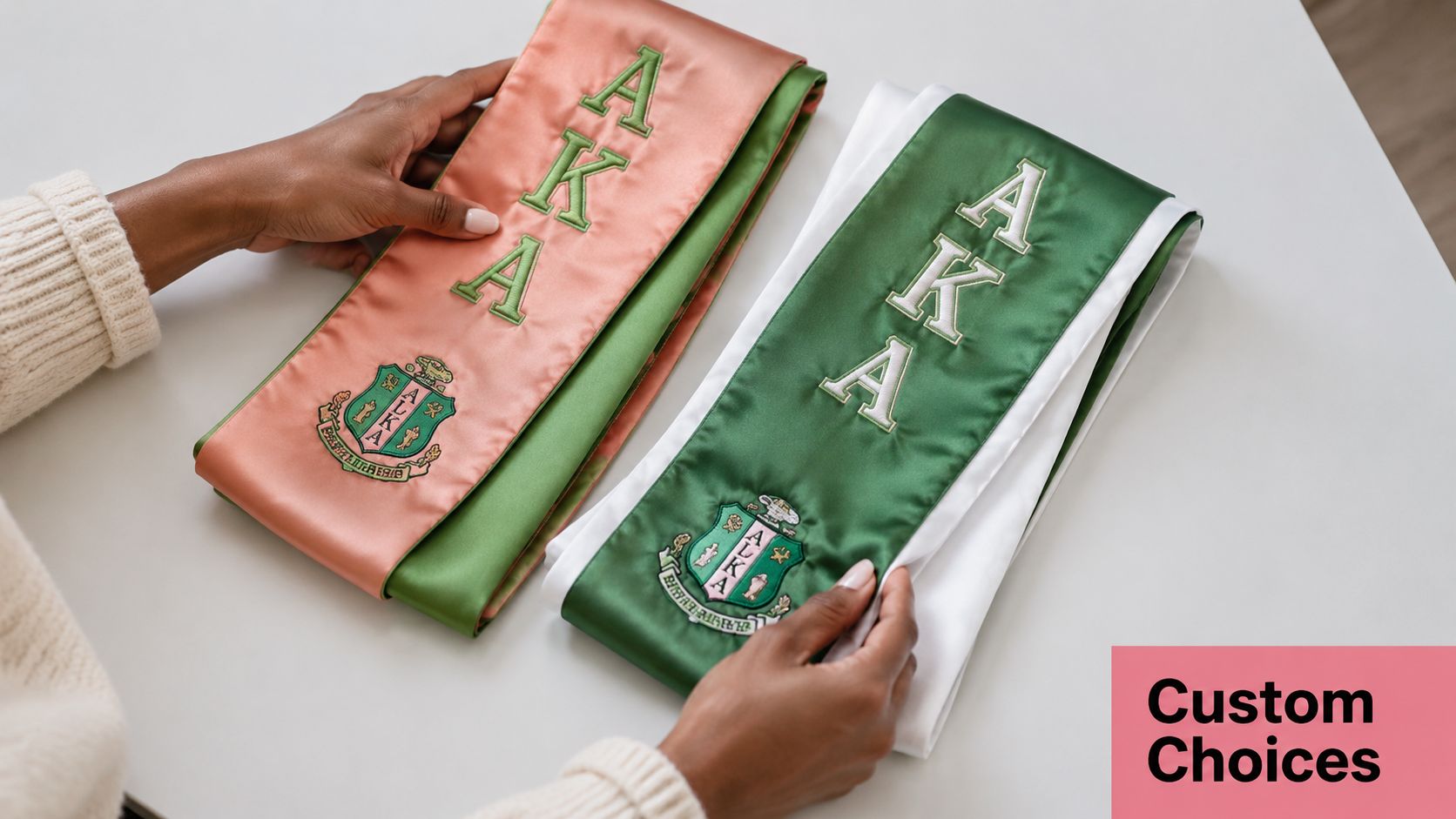 A person displays two custom AKA graduation stoles in pink and green colors on a white table.