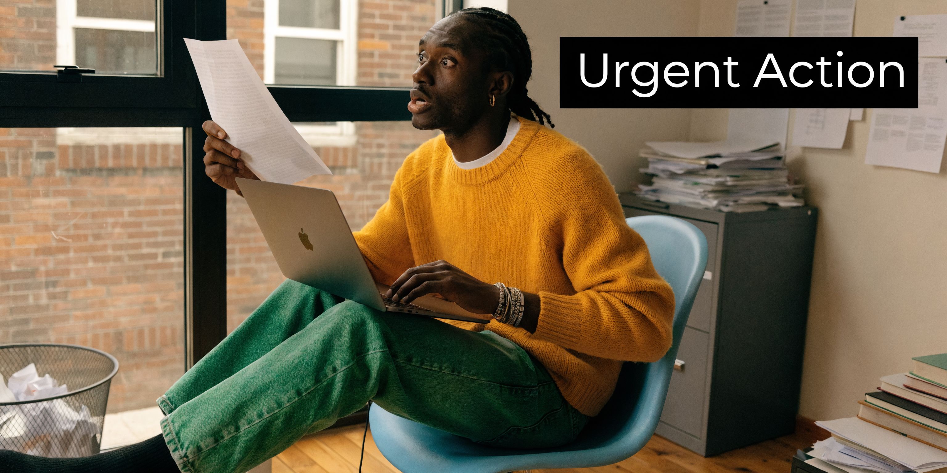 A concerned man in a yellow sweater holds a document while looking shocked at his laptop screen.