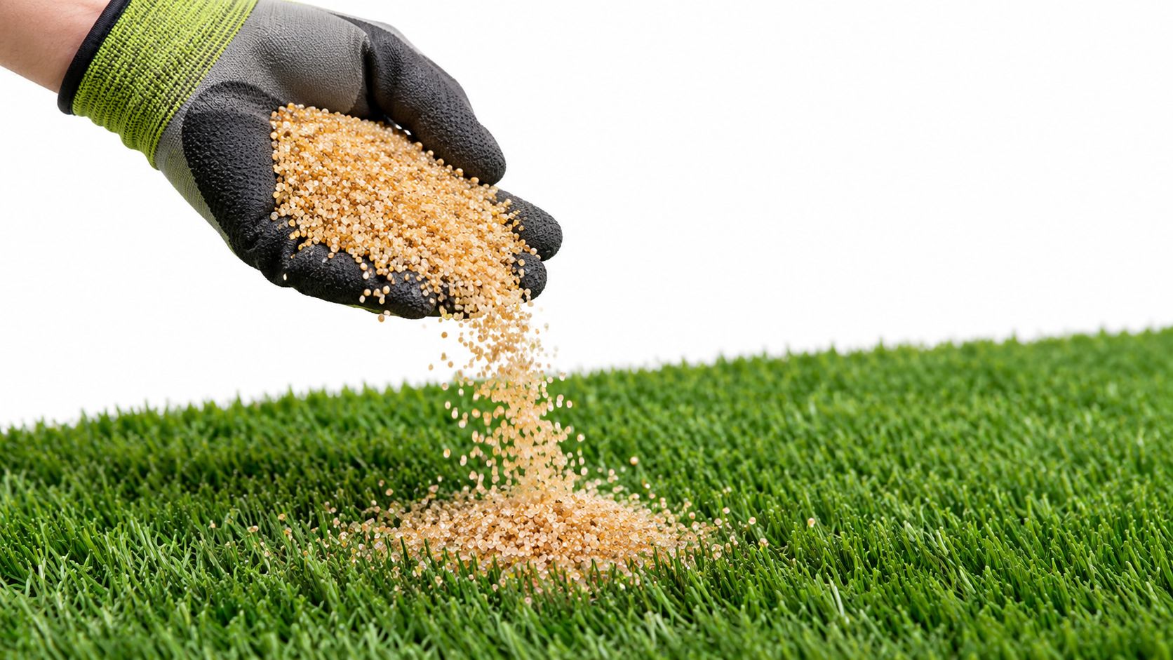 A gloved hand spreading small, light brown artificial turf infill pellets onto a patch of green grass.