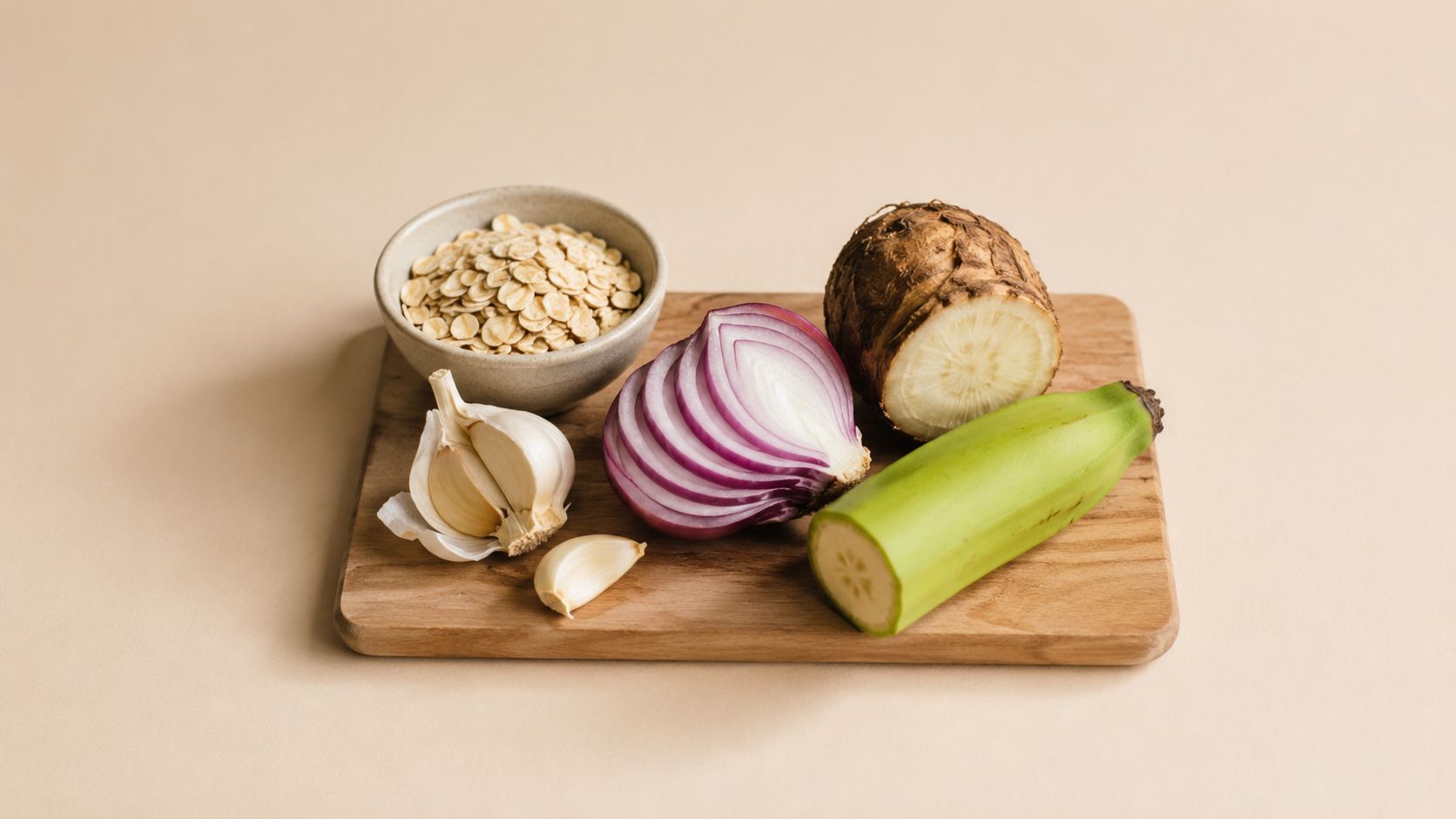 A wooden cutting board with a bowl of oats, sliced red onion, garlic cloves, taro, and plantain.