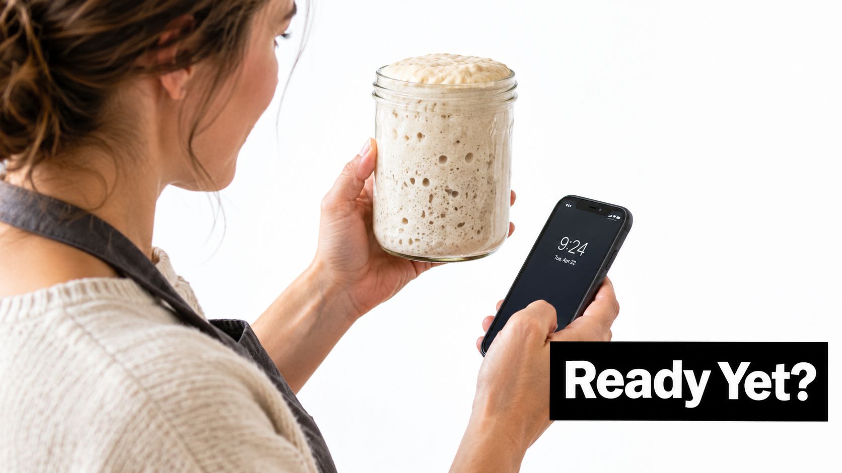 A woman holding a jar of bubbly sourdough starter while checking the time on her smartphone.