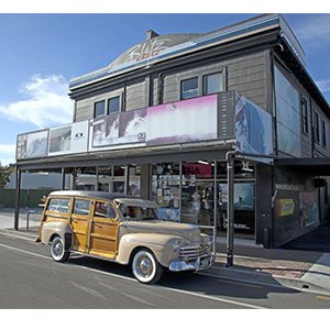 A vintage beige wood-paneled station wagon, a "woody car," parked in front of a surf shop.