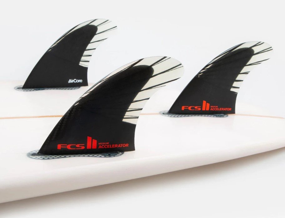A close-up view of three FCS II surfboard fins mounted on the tail of a white surfboard.