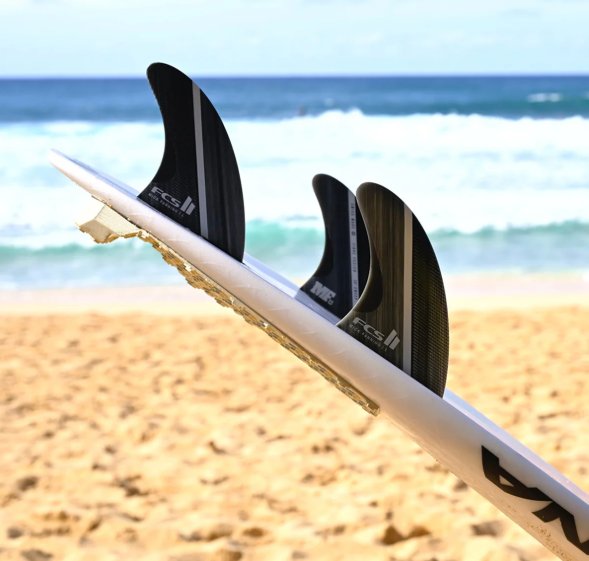 A close-up view of surfboard fins attached to a white surfboard on a sandy beach.