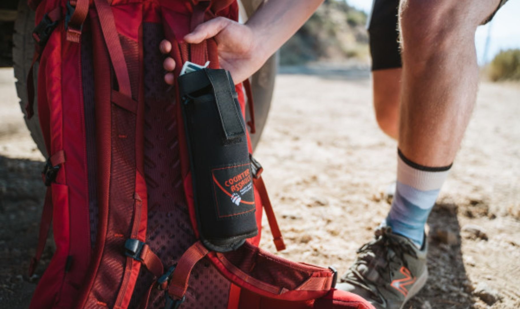 A person's hand holds a black bear spray holster attached to a red backpack on a dirt path.