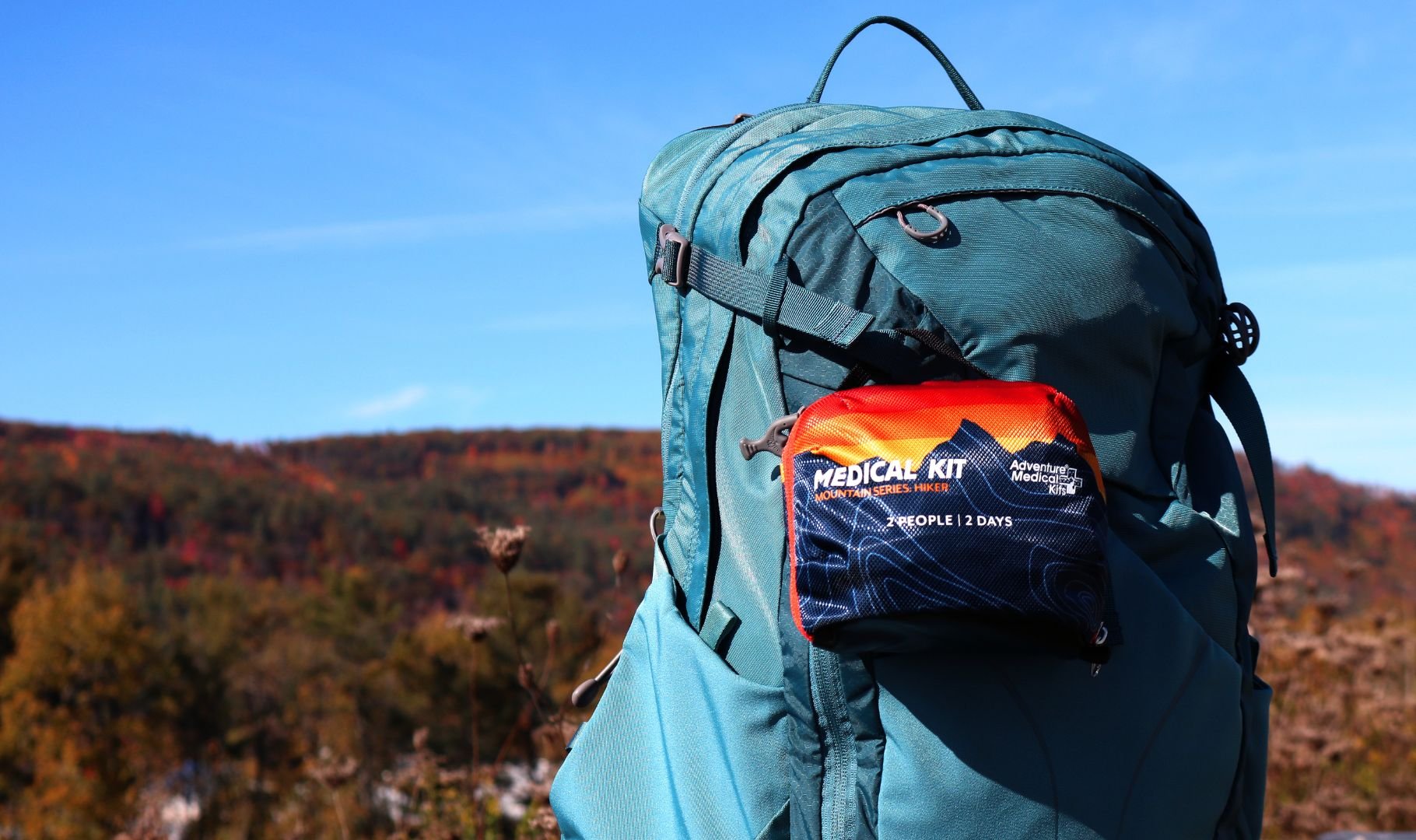 A blue hiking backpack with an attached red medical kit in a scenic autumn landscape.