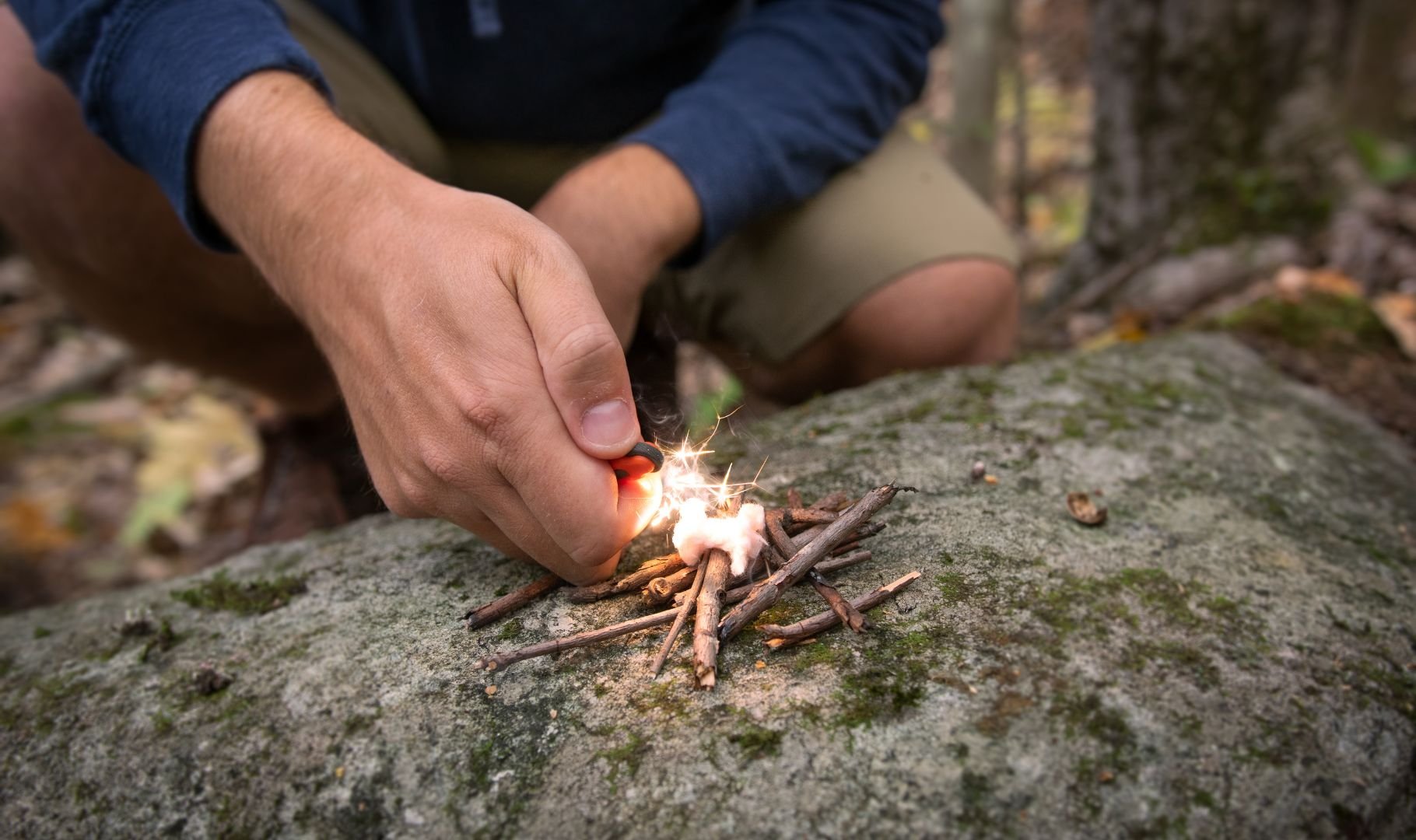 A person crouches, using a fire starter to ignite a small pile of twigs and cotton on a mossy rock.