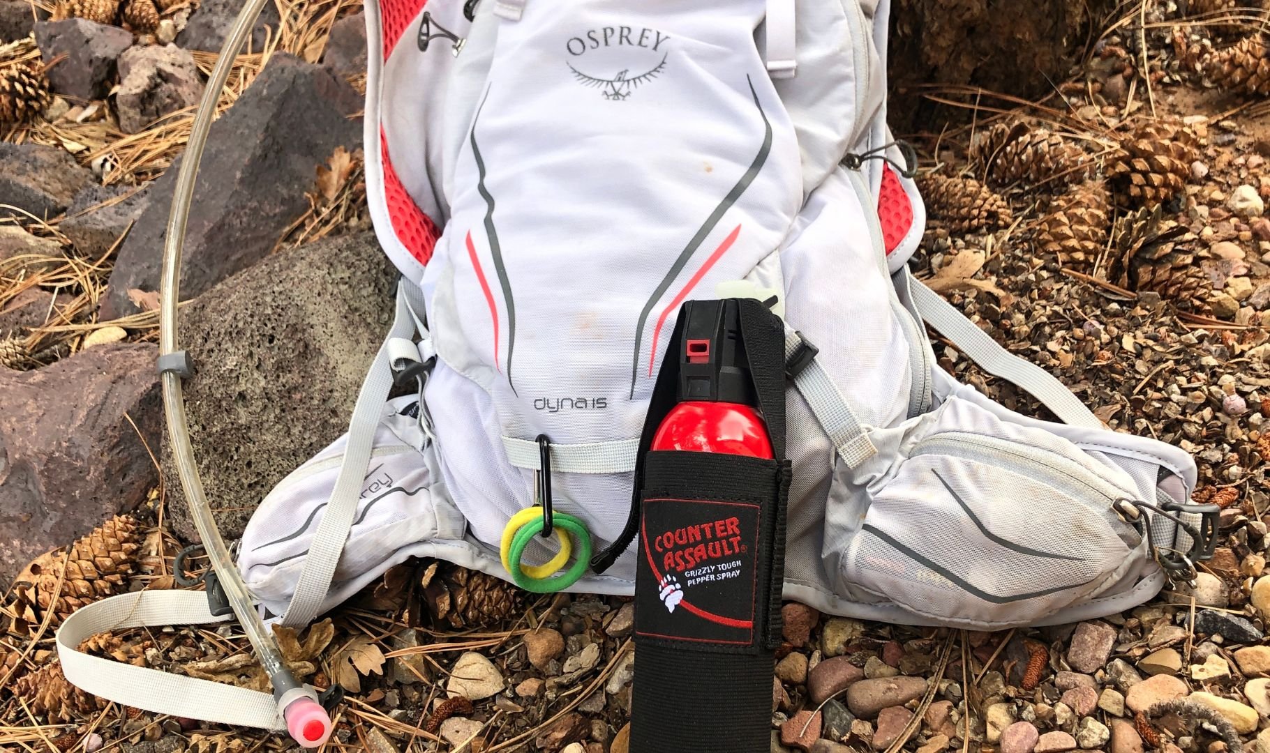 A white Osprey backpack with a hydration tube and red bear spray, resting on a rocky forest floor.