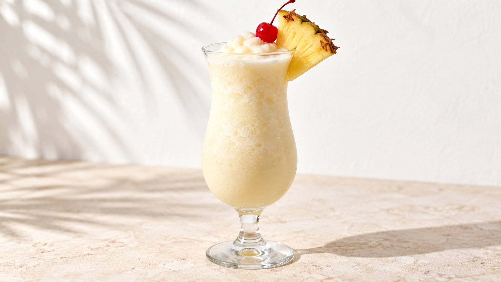 A frosty piña colada cocktail in a glass, garnished with a pineapple slice and cherry, under palm shadows.