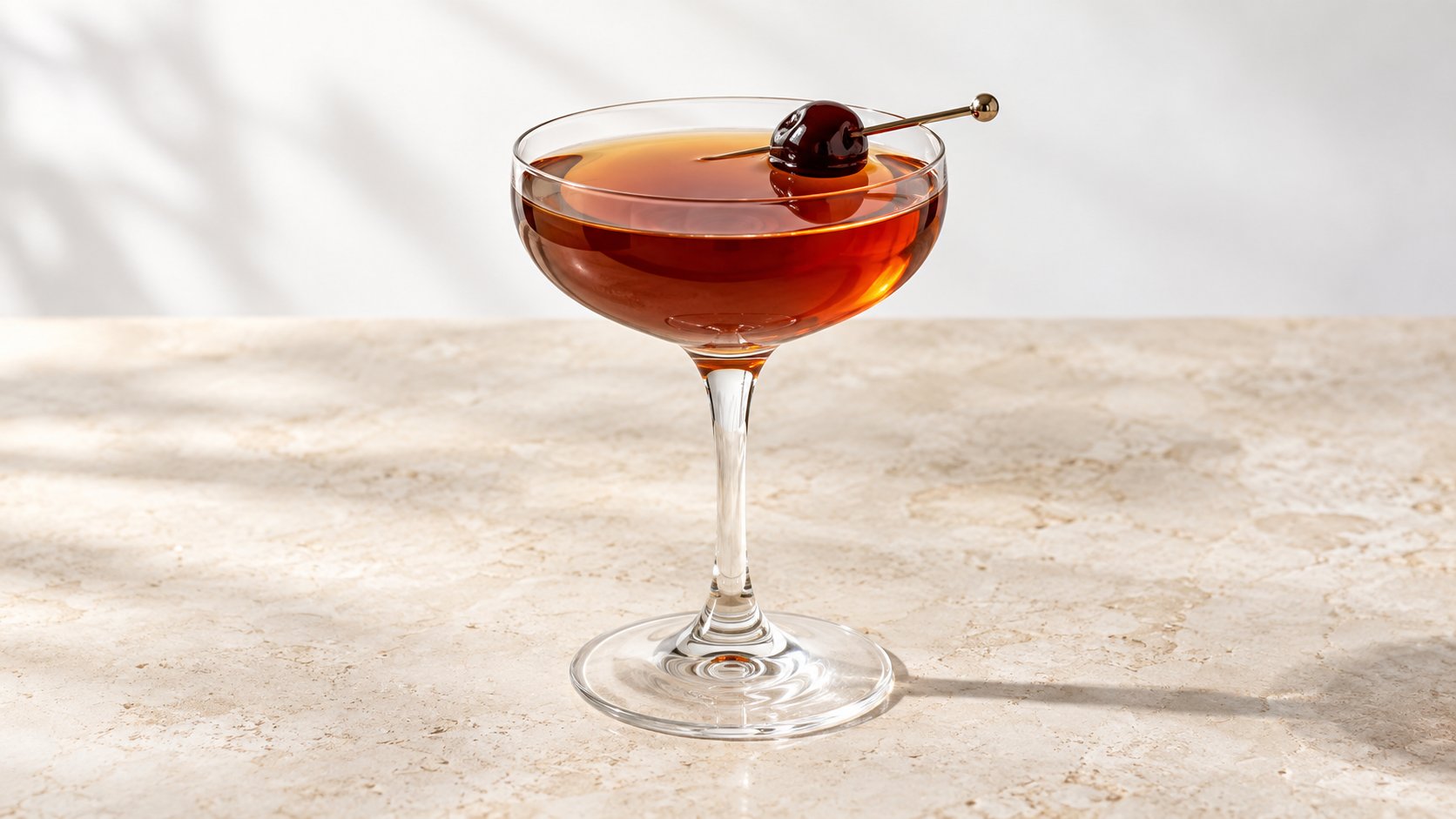 A sophisticated brown cocktail in a coupe glass, garnished with a cherry, on a textured table.