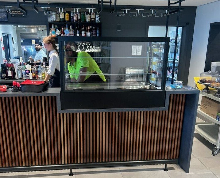 Two staff members working behind a cafe bar with bottles, a display case, and a kitchen area.