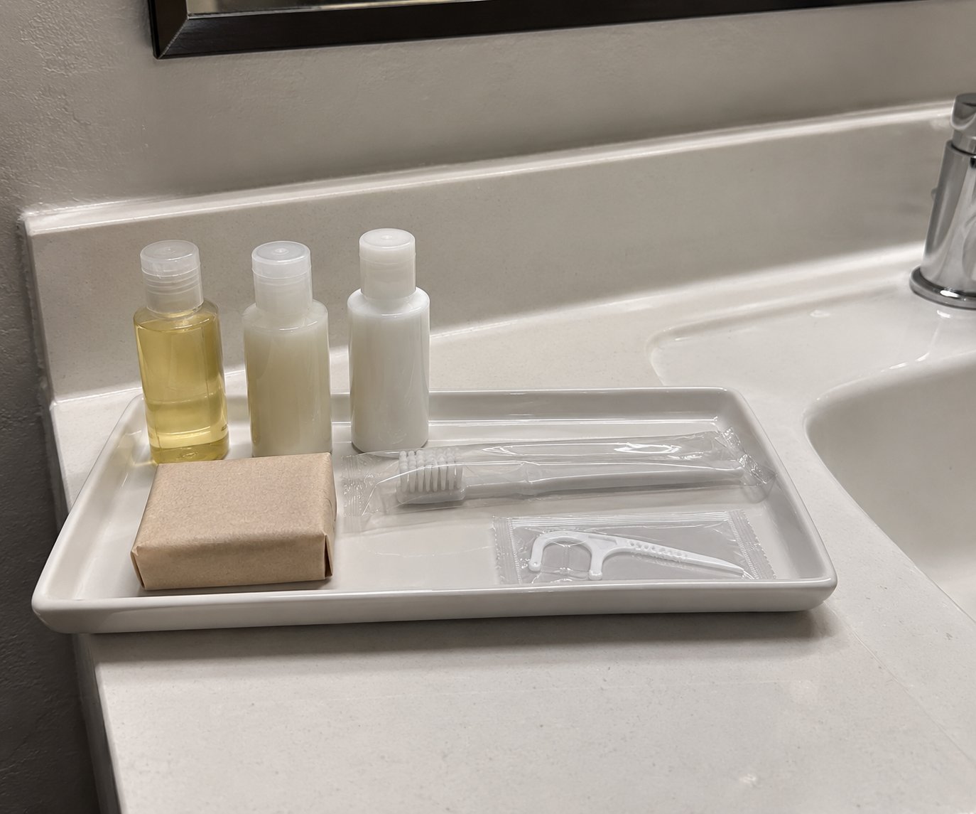 A white tray holds travel-size toiletries: shampoo, conditioner, lotion, soap, toothbrush, and floss, on a bathroom counter.