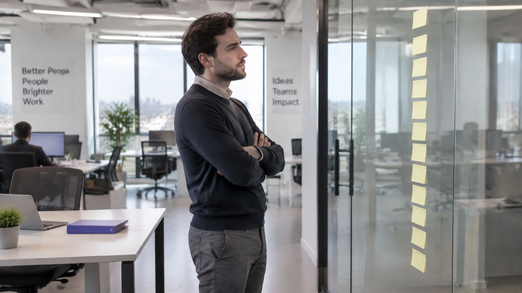 A professional man in an office standing with crossed arms looking at sticky notes on glass wall.