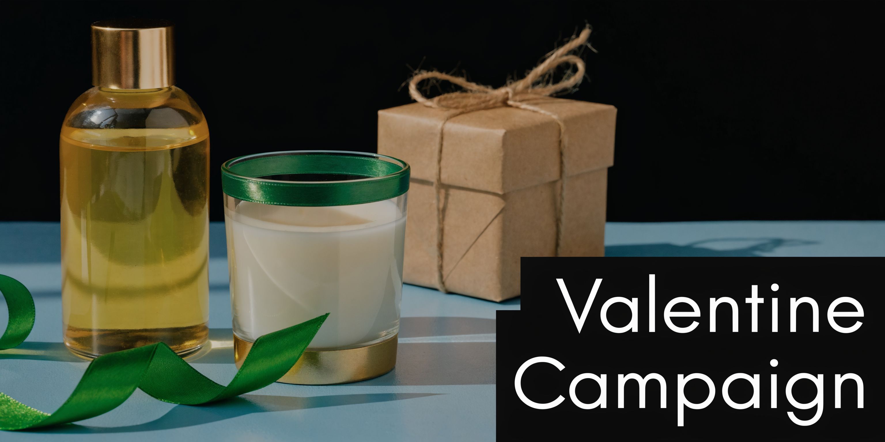 A glass candle with green ribbon, a bottle of oil, and a wrapped gift on blue surface.