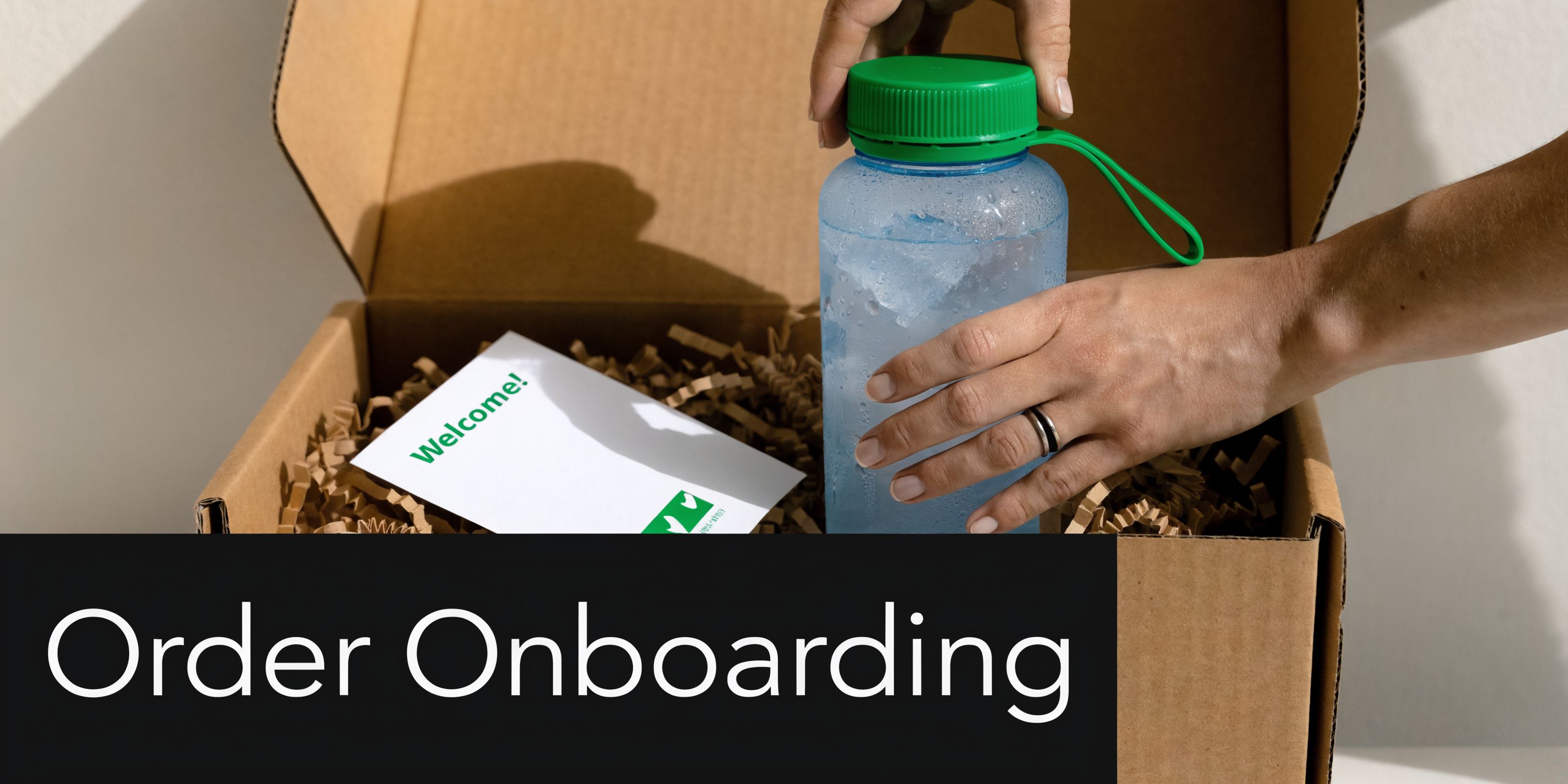 A person placing a blue water bottle into a cardboard box filled with paper filler and a welcome note.