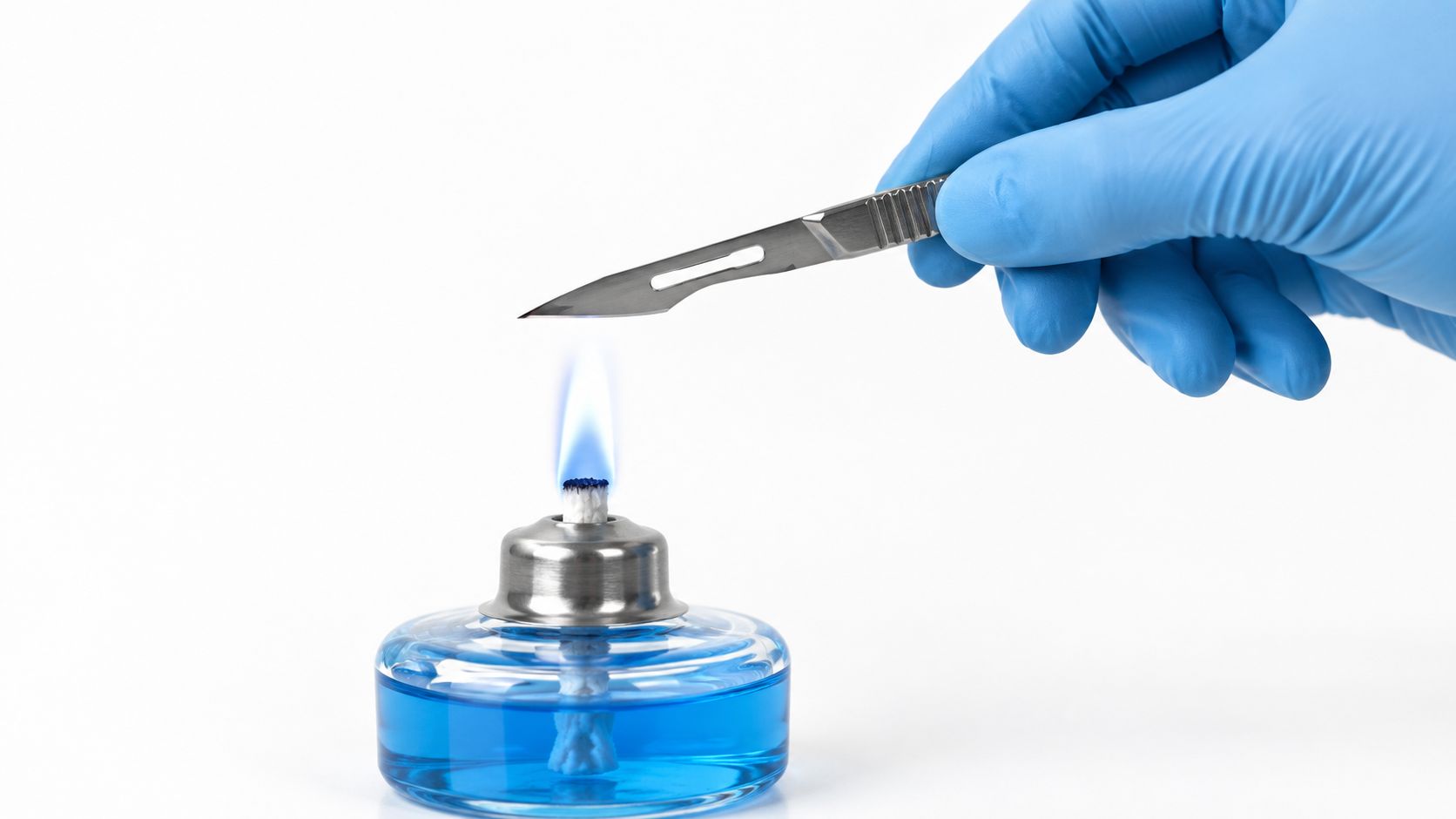 A gloved hand holds a stainless steel scalpel blade over the blue flame of a laboratory alcohol lamp.