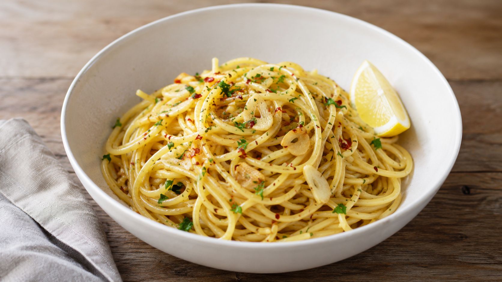 A close-up of a white bowl filled with spaghetti aglio e olio topped with parsley and lemon.