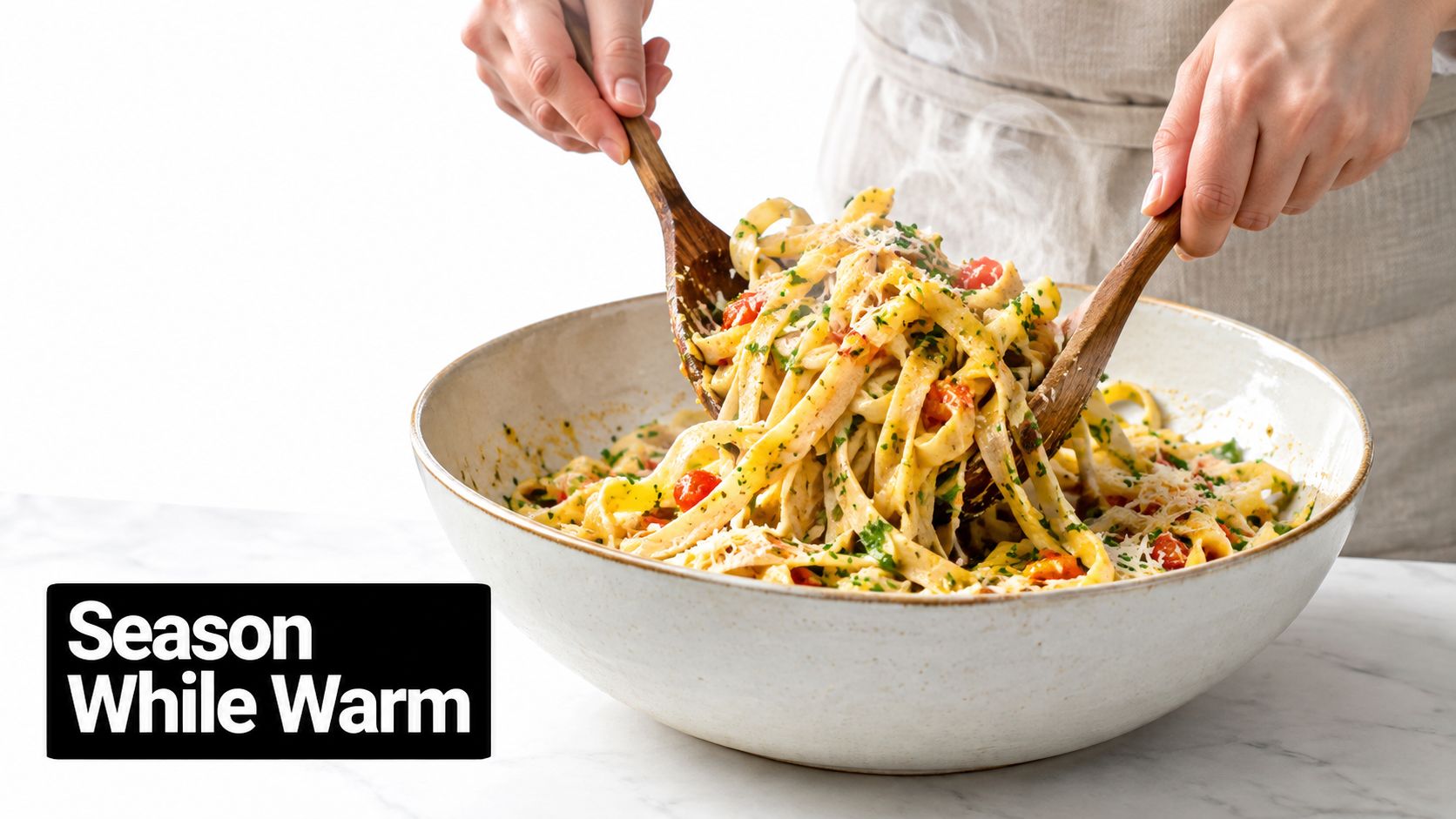 A person mixing freshly cooked fettuccine pasta with tomatoes and herbs using wooden spoons in a bowl.
