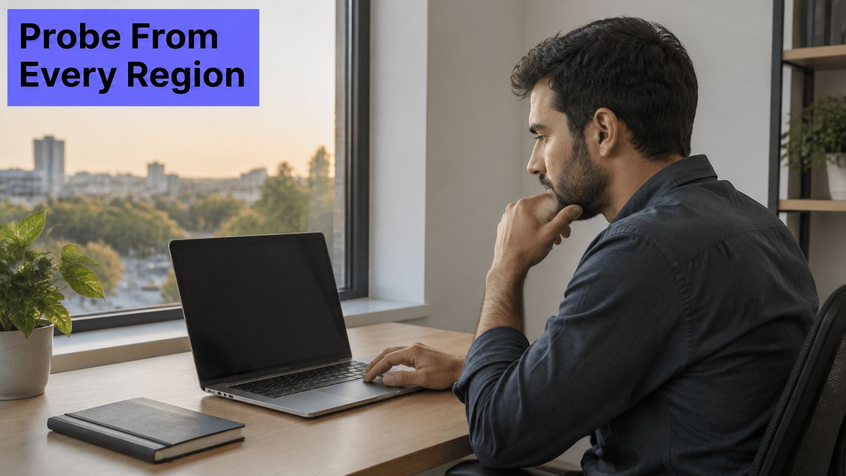 A man working on his laptop while sitting at a desk by a window with a city view.