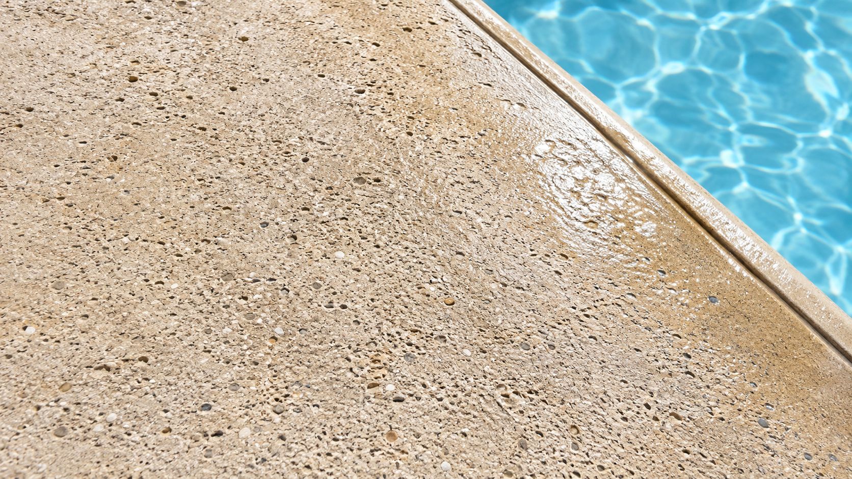 A close-up view of a textured concrete pool deck beside a sparkling blue swimming pool water.