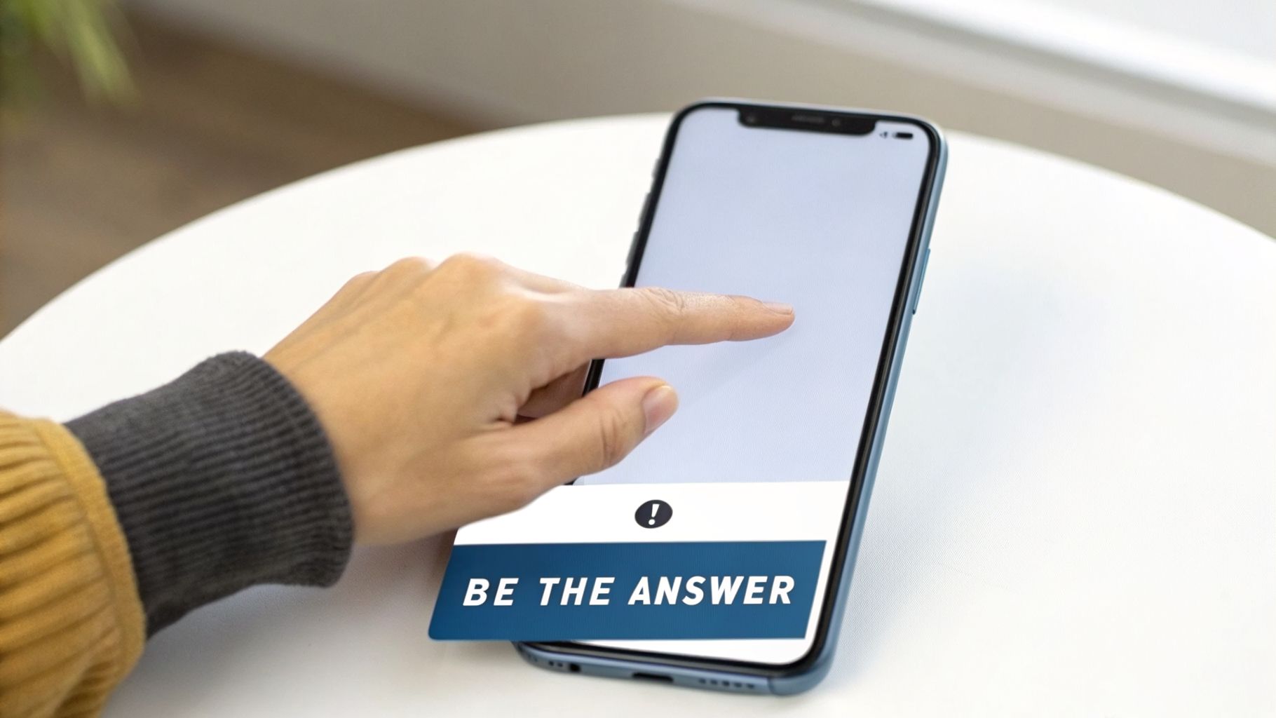 A person's hand taps a smartphone screen displaying 'BE THE ANSWER' on a white table.