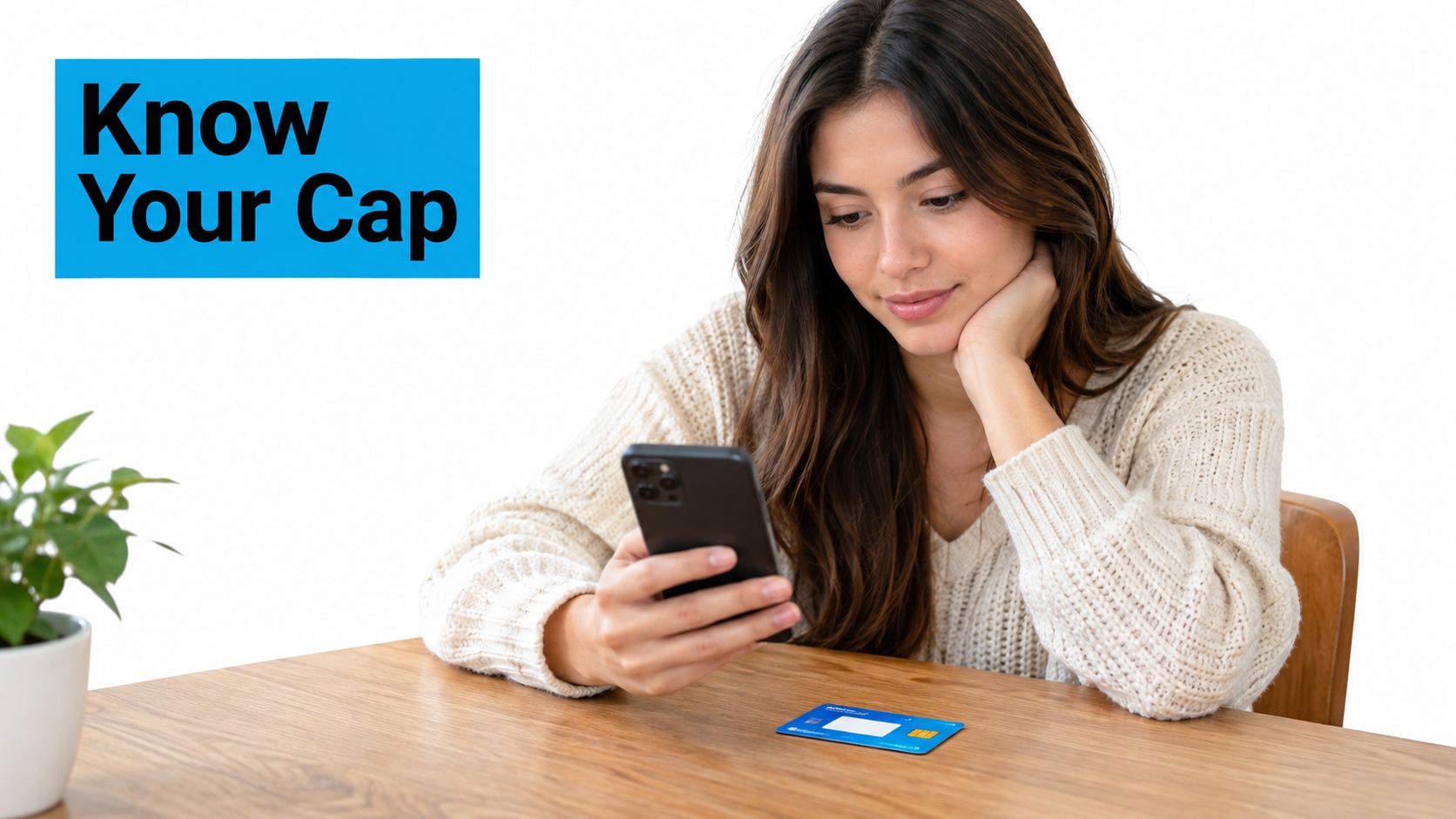 A young woman sits at a wooden table looking at her smartphone with a bank card nearby.