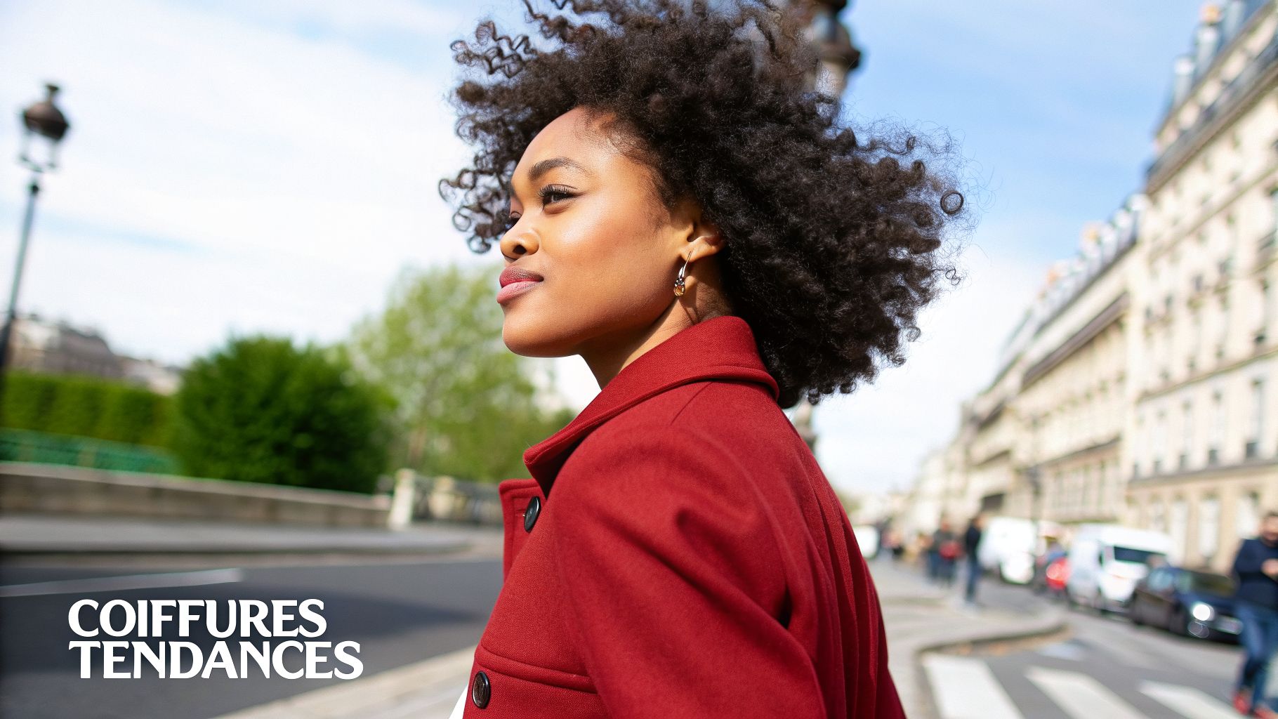 Une femme souriante aux cheveux afro bouclés, vêtue d'un manteau rouge, marchant dans une rue de ville ensoleillée.