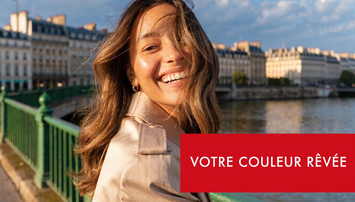 Une femme souriante aux cheveux châtains brillants pose sur un pont de Paris devant la Seine.