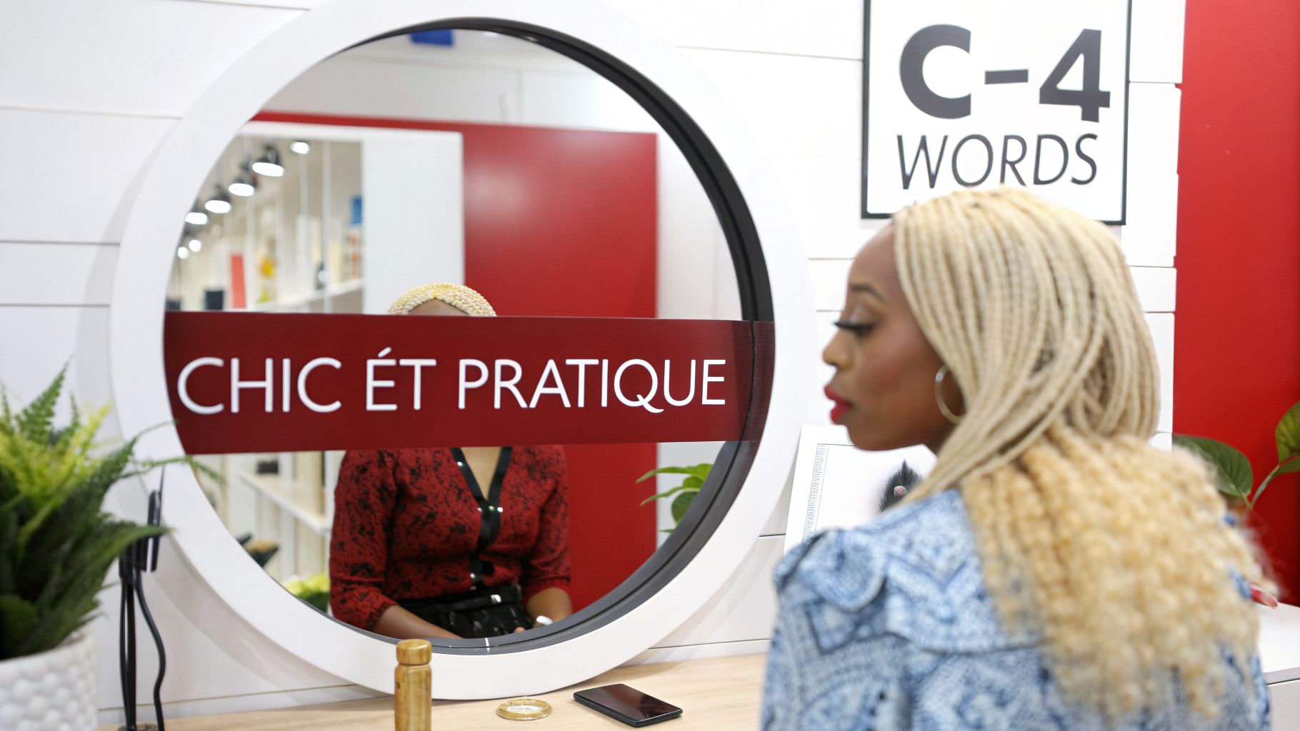 Une femme aux tresses blondes, vêtue de bleu, contemple son reflet dans un miroir circulaire affichant "CHIC ÉT PRATIQUE".