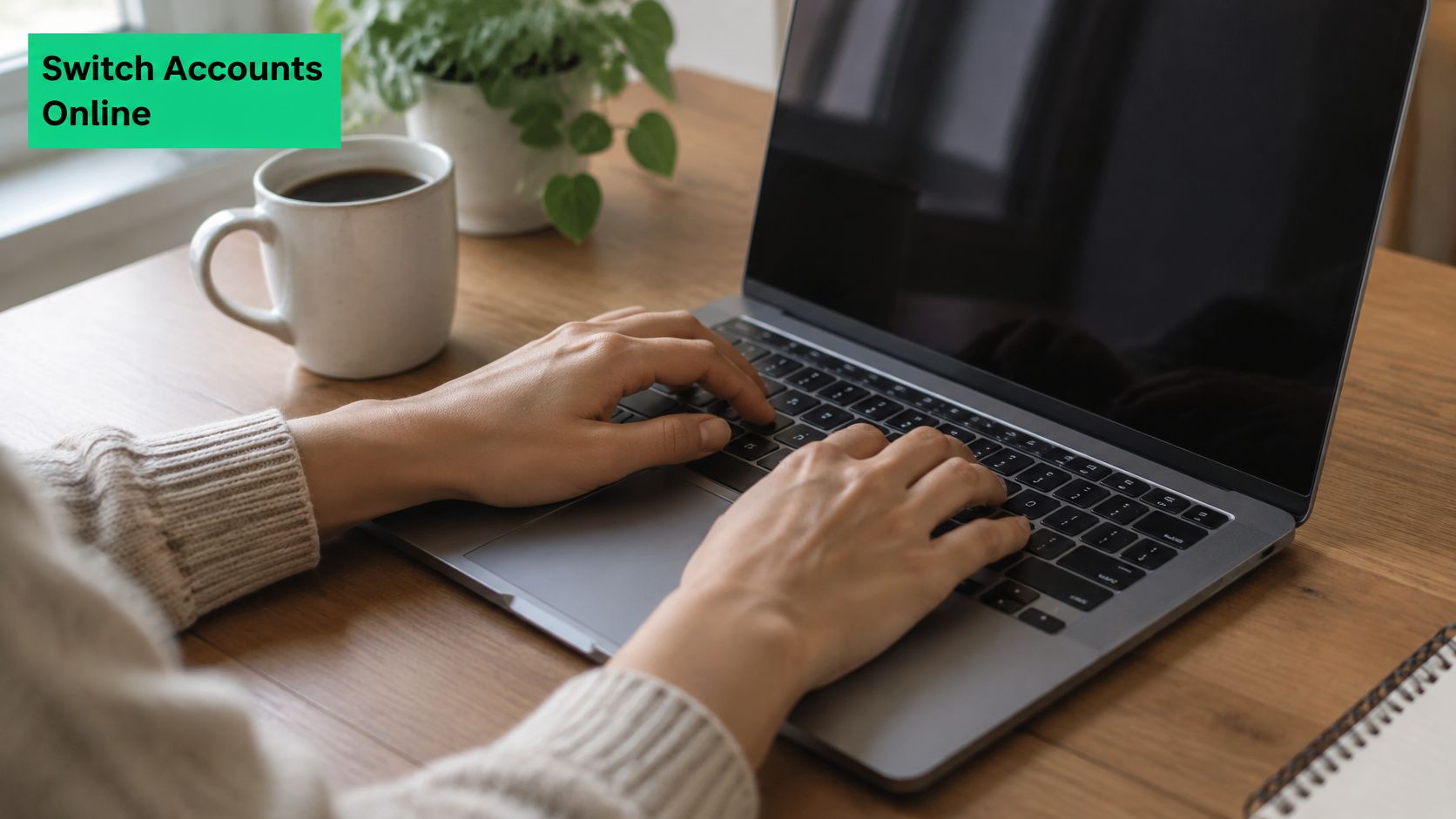 A person typing on a laptop at a wooden desk with a coffee mug and plant nearby.