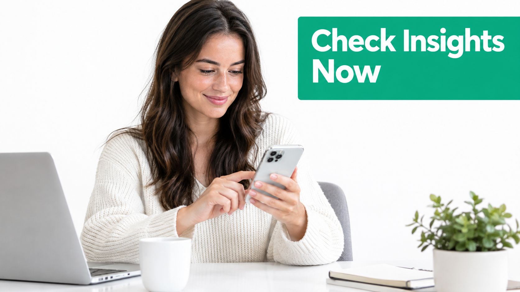 A woman smiling while looking at her smartphone with a laptop and plant on her desk